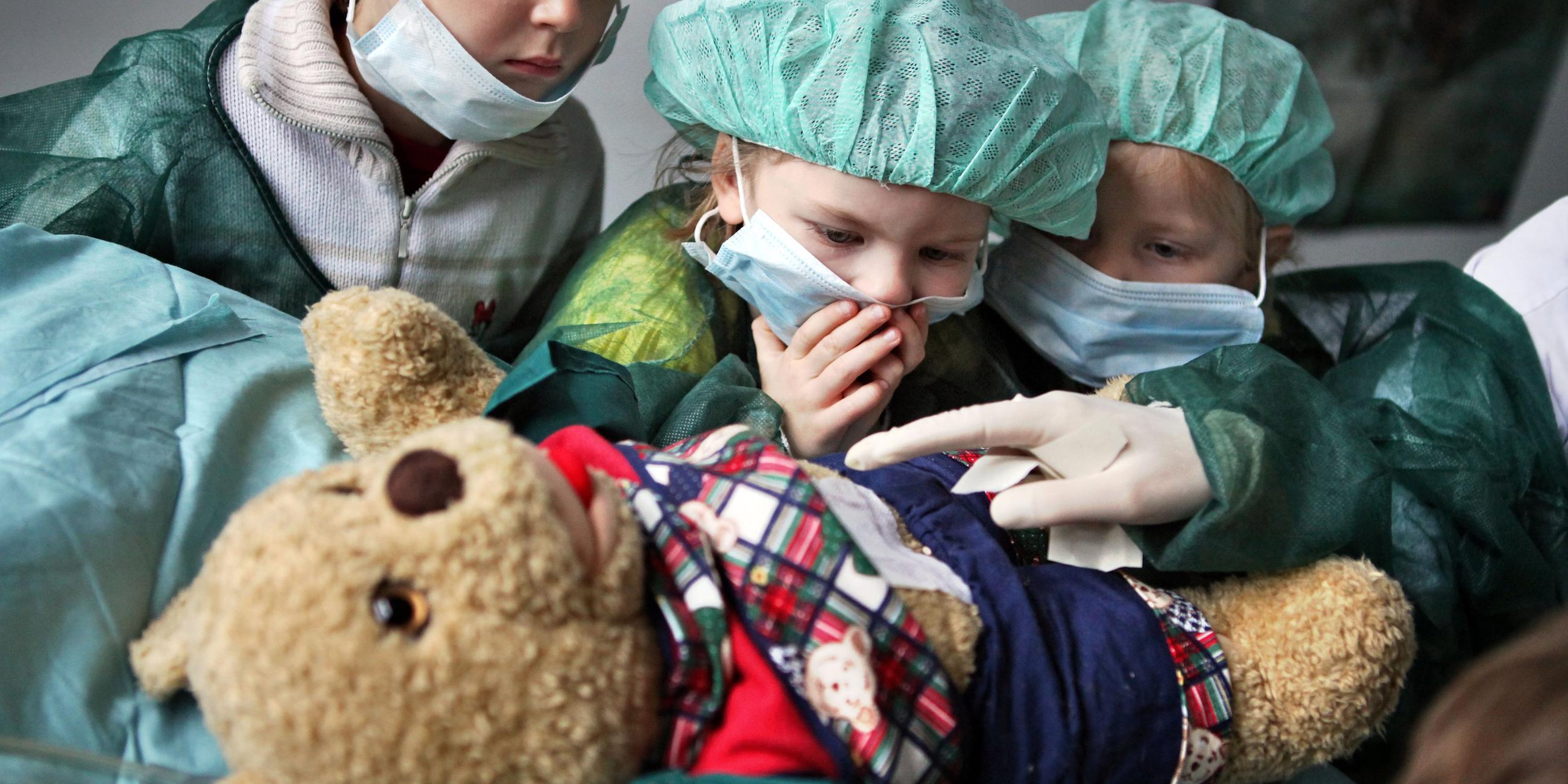 Drei Kindergartenkinder in klinischer Schutzkleidung und mit Maske sehen dabei zu, wie einem Teddybär im Vordergrund spielerisch der Blinddarm entfernt wird.