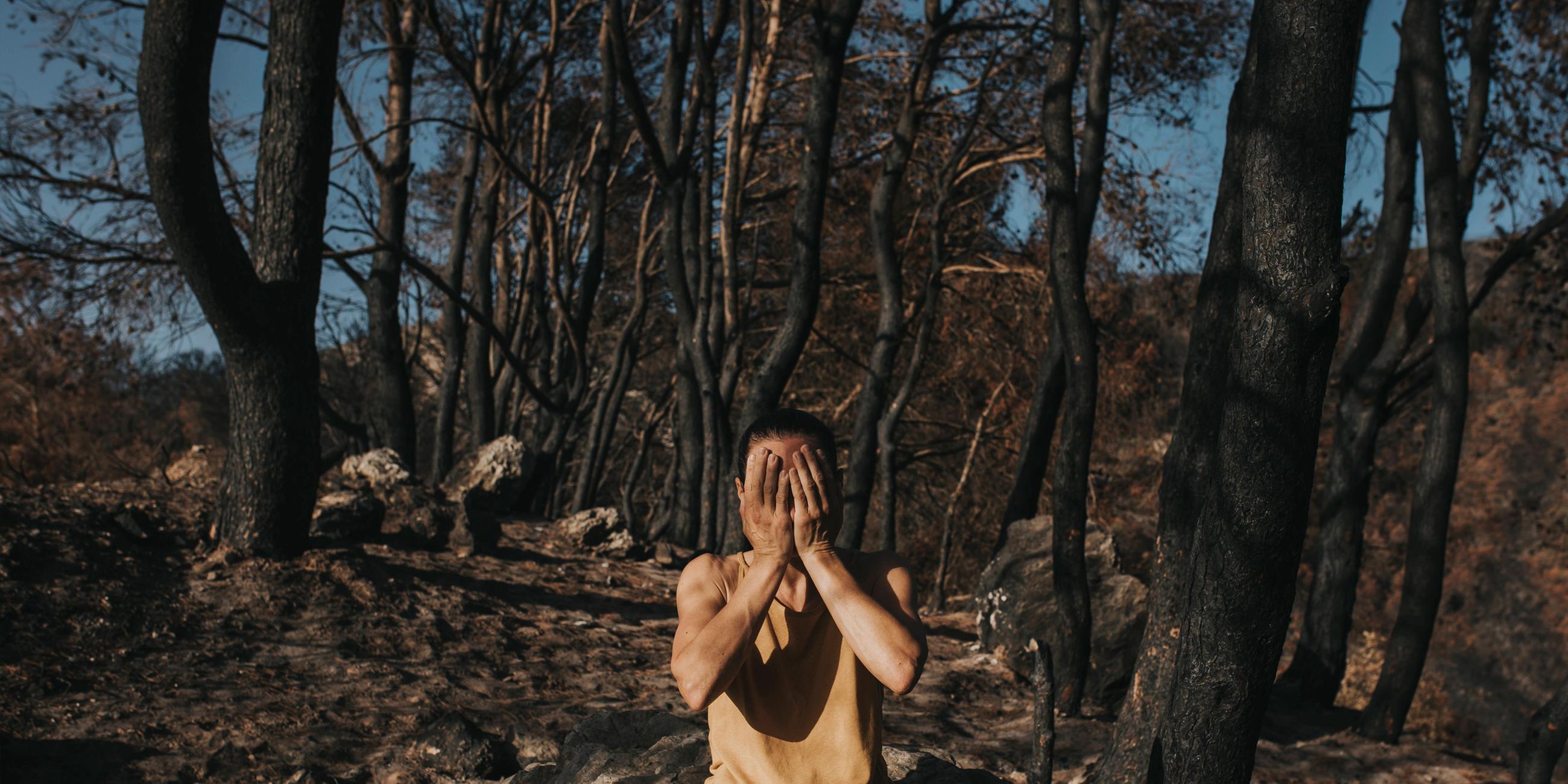 Symbolbild: Eine verzweifelte Frau, die ihr Gesicht verdeckt und auf einem Felsen in einem verbrannten Wald sitzt.