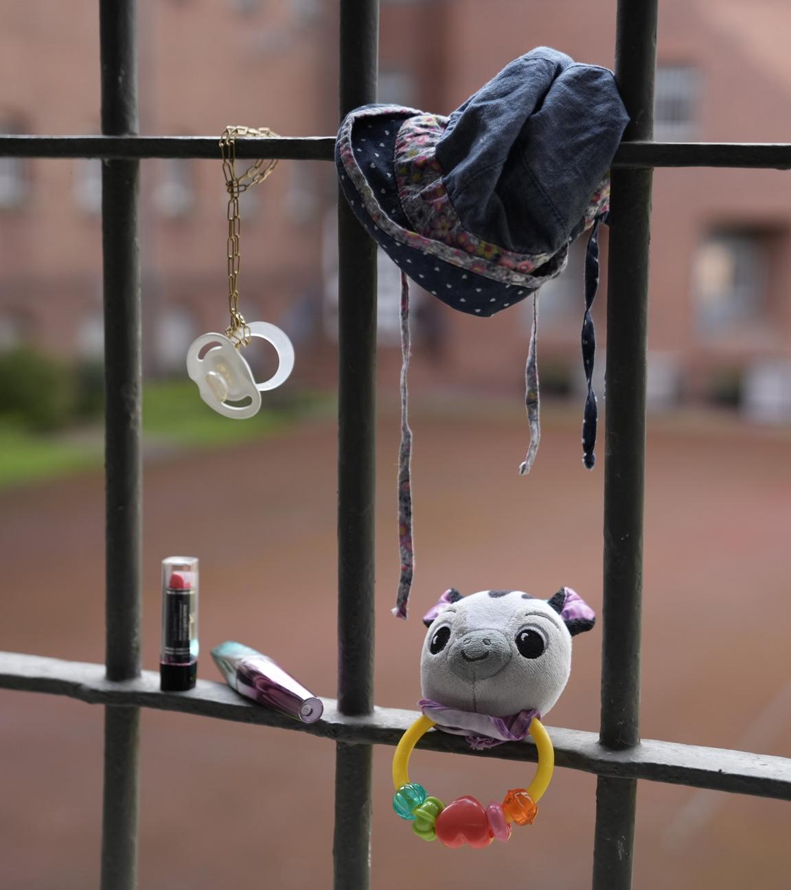 An einem Gefängnisgitter hängen ein Schnuller, eine Babymütze und eine Rassel. Auf einer Gitterstrebe steht ein Lippenstift.