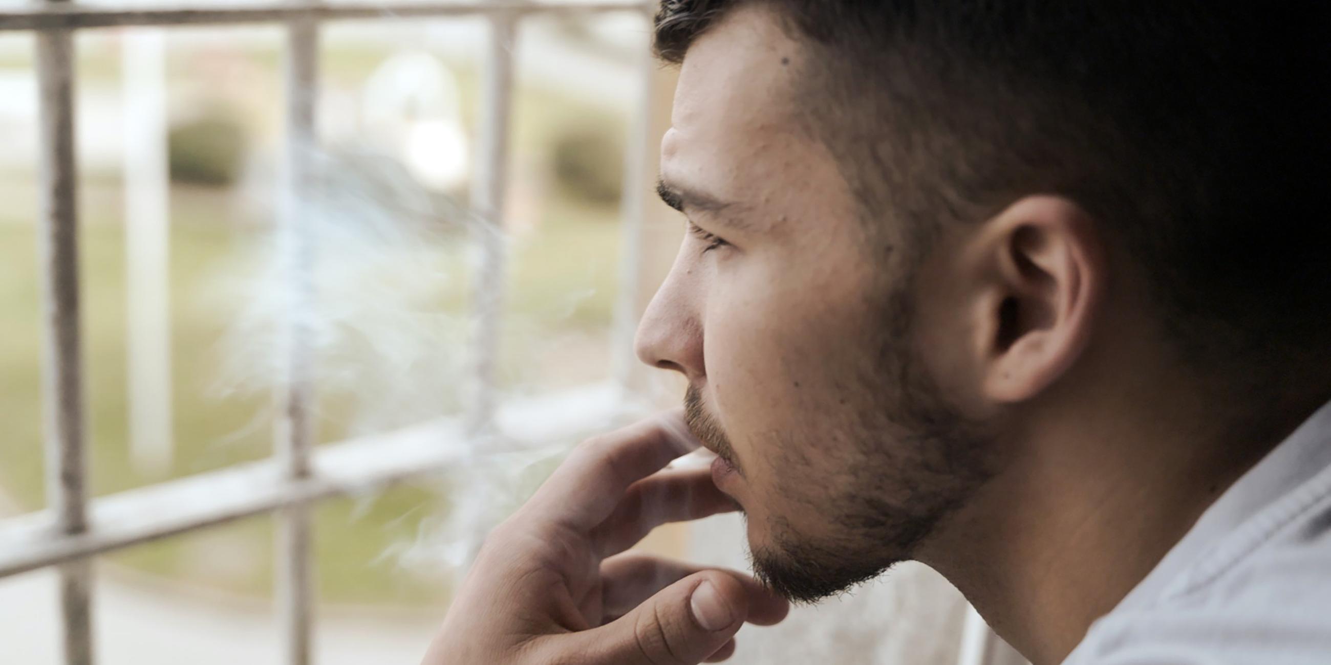 Ein junger Mann steht an einem Gitterfenster und raucht. Er blickt nach draußen.