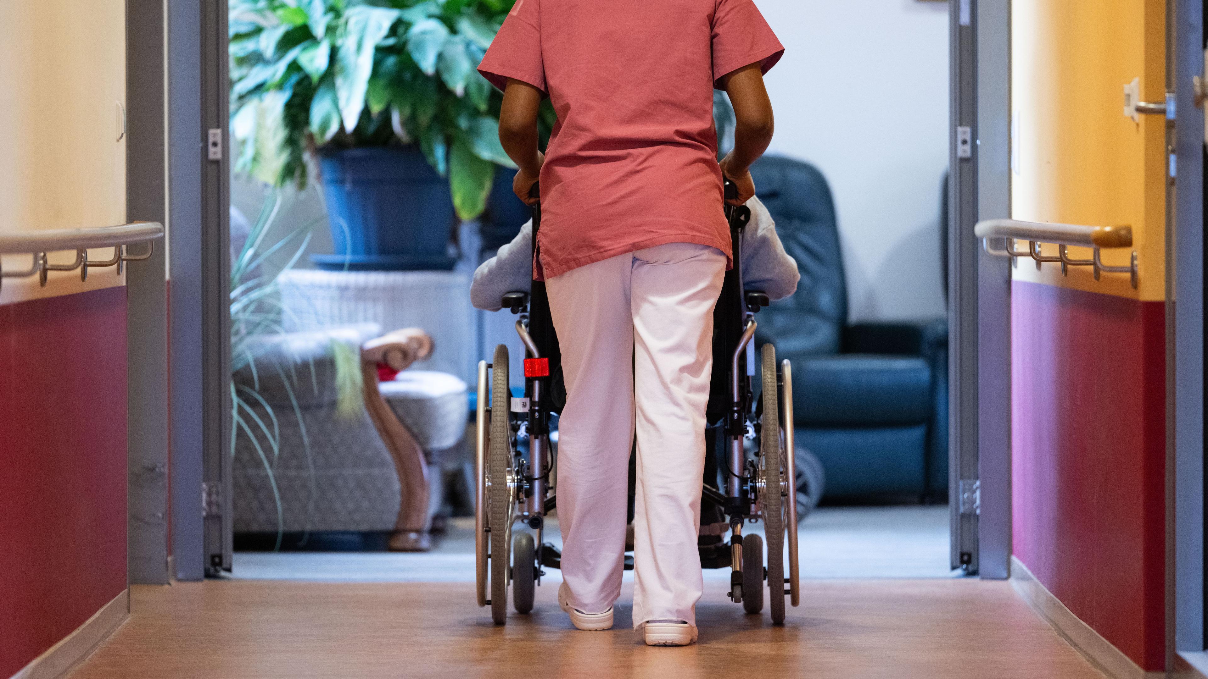 Symbolbild: Frau schiebt Mensch im Rollstuhl auf Pflegeheim-Gang