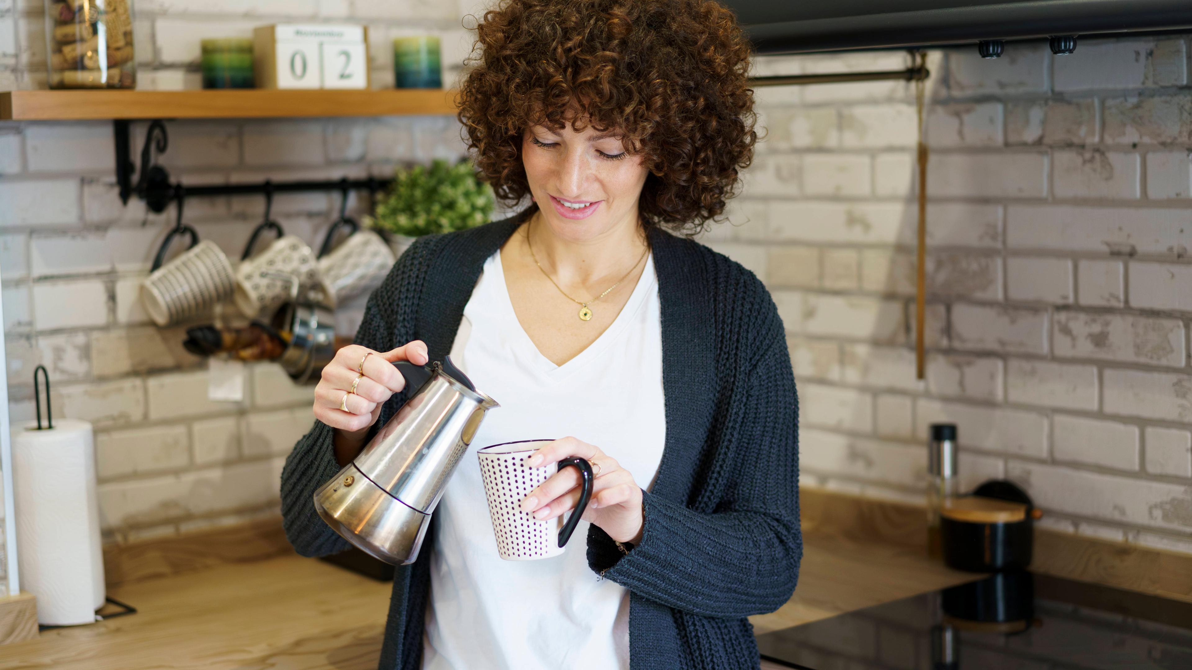 Eine Frau schenkt sich in der Küche am Tresen ein Tasse Kaffee aus einer Kanne ein.