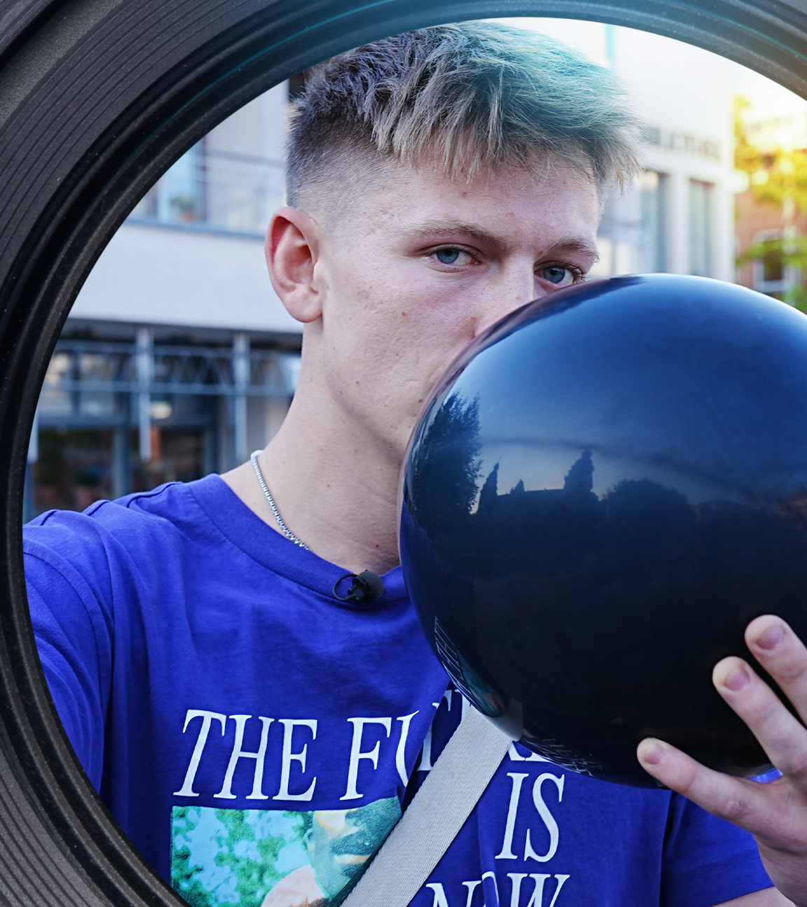 Ein junger Mann hält sich mit einer Hand einen Luftballon vor den Mund. Mit der anderen Hand streckt er eine Kartusche Lachgas in die Kamera.