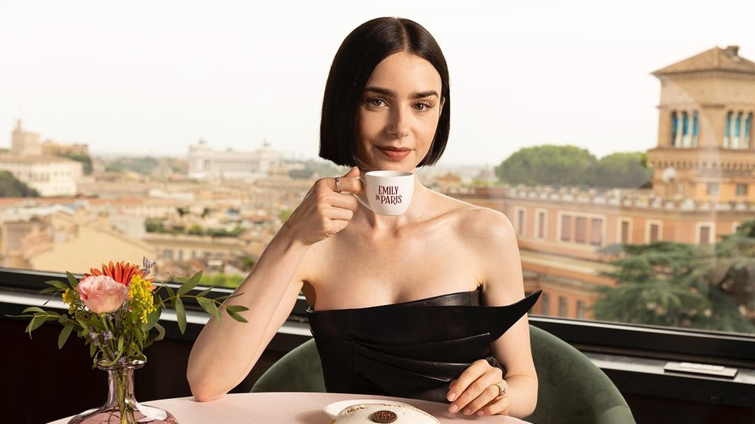 Die Schauspielerin Lily Collins hält eine Tasse mit dem Schriftzug "Emily in Paris". Im Hintergrund ist die Stadt Rom zu sehen.