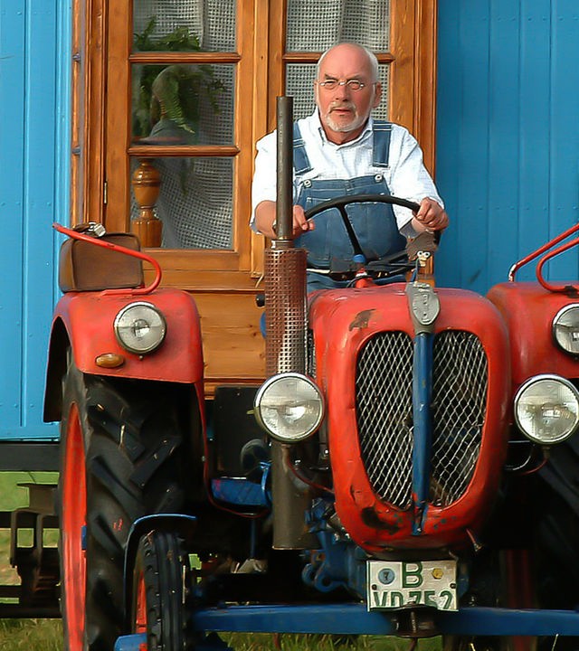 Peter Lustig sitzt auf einem Traktor, der den Bauwagen über eine Wiese zieht