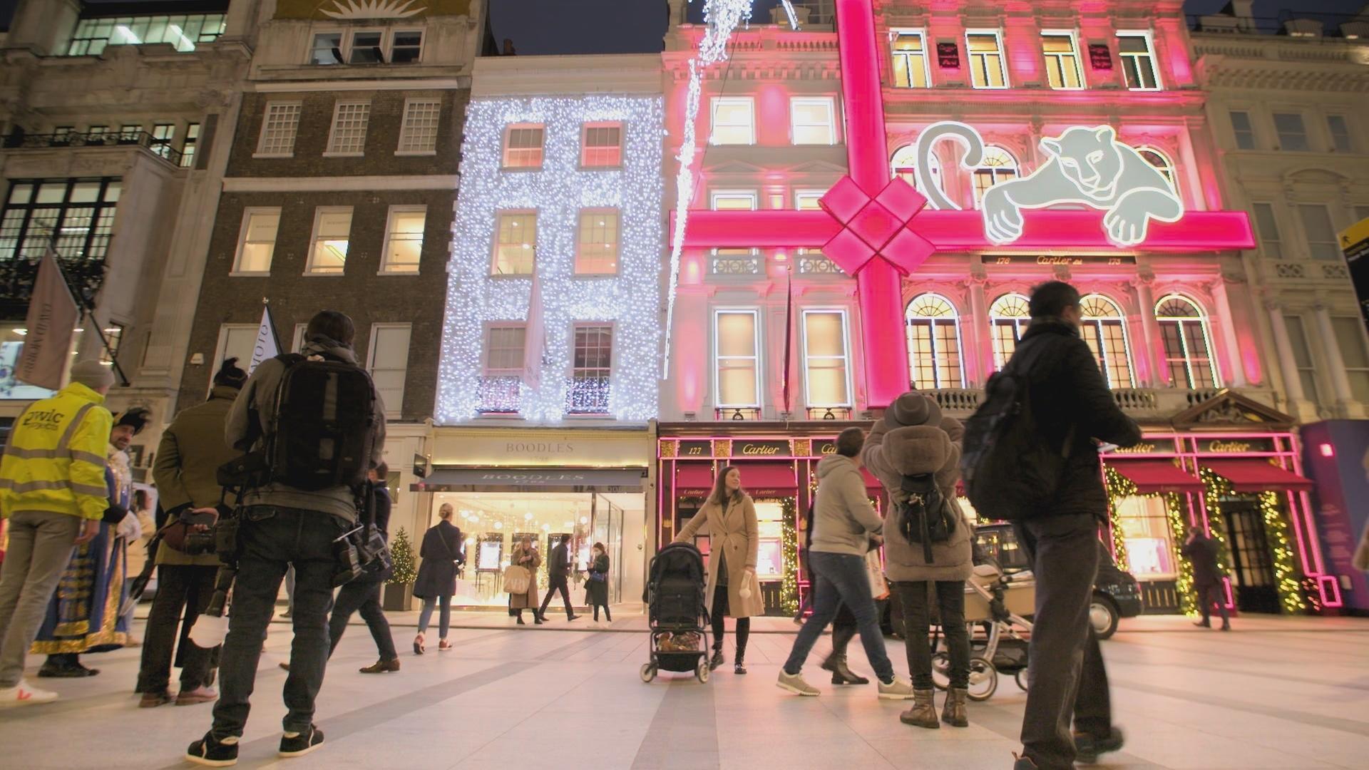 Zahlreiche Fußgänger in winterlicher Kleidung vor einer weihnachtlich beleuchteten Häuserfront. Im Erdgeschoss der Häuser sind Geschäfte der Marken Boodles und Cartier zu erkennen.