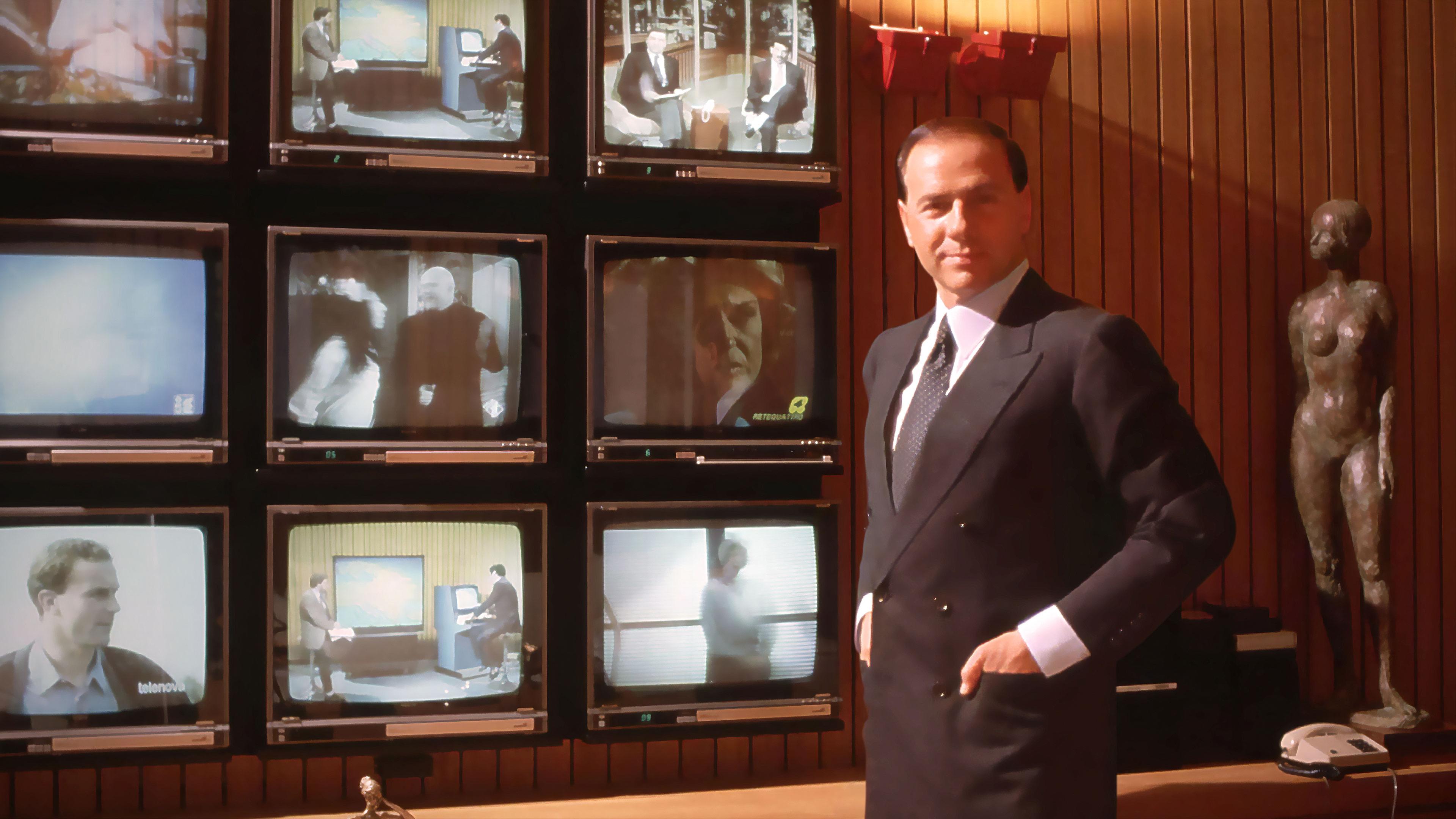 Silvio Berlusconi steht vor einer Wand mit mehreren Fernsehern.
