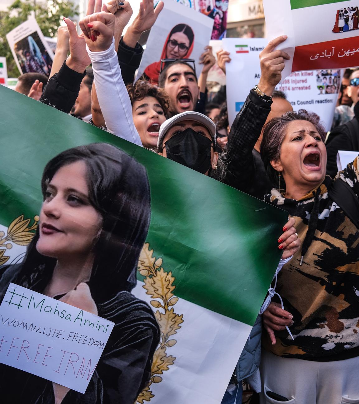 Iranerinnen und Iraner halten Bilder von Mahsa Amini bei einem Protest hoch. Ihre Hände sind rot bemalt.