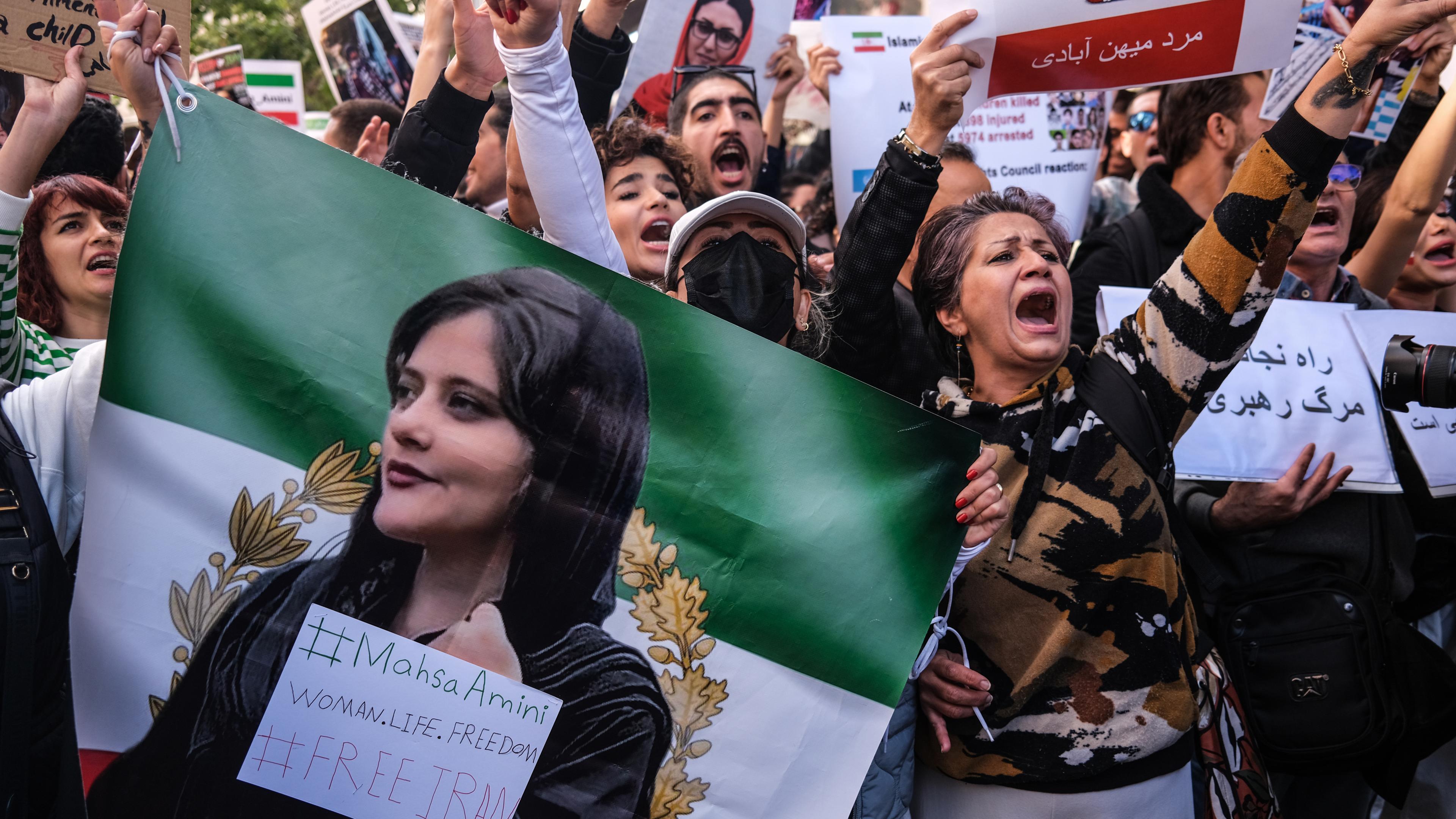 Iranerinnen und Iraner halten Bilder von Mahsa Amini bei einem Protest hoch. Ihre Hände sind rot bemalt.