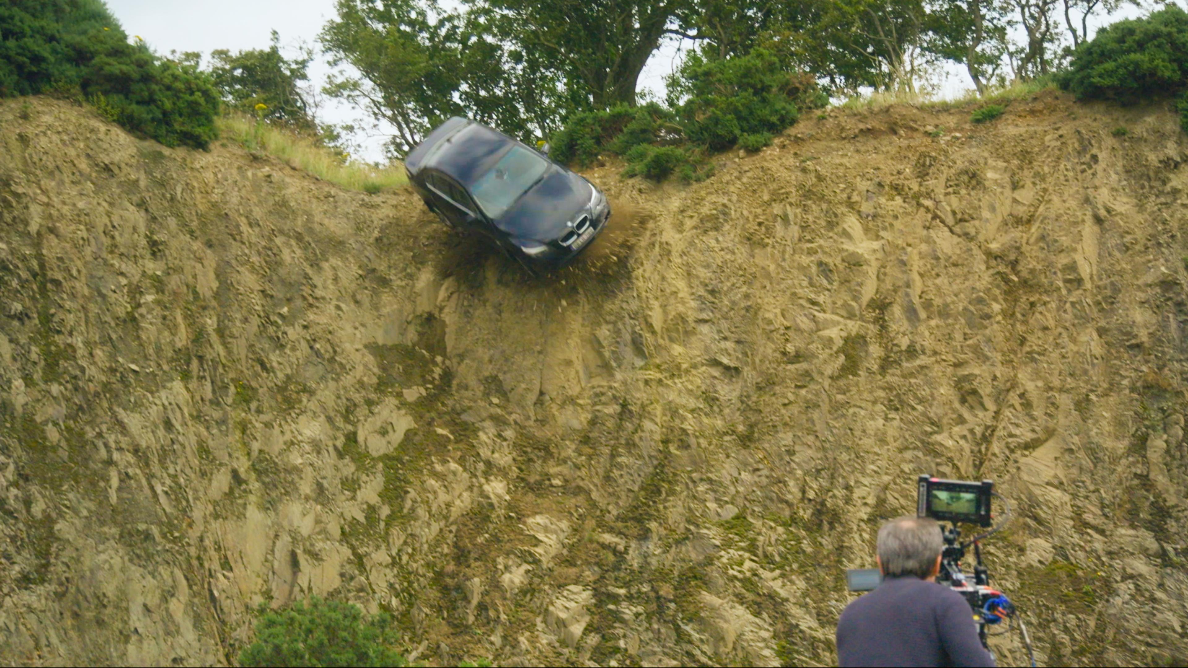 "The Tourist - Irisches Blut: Making-of": Eine dunkle BMW-Limousine stürzt eine Klippe herab. Die Szene wird von einem Kameramann rechts unten im Bild aufgenommen.