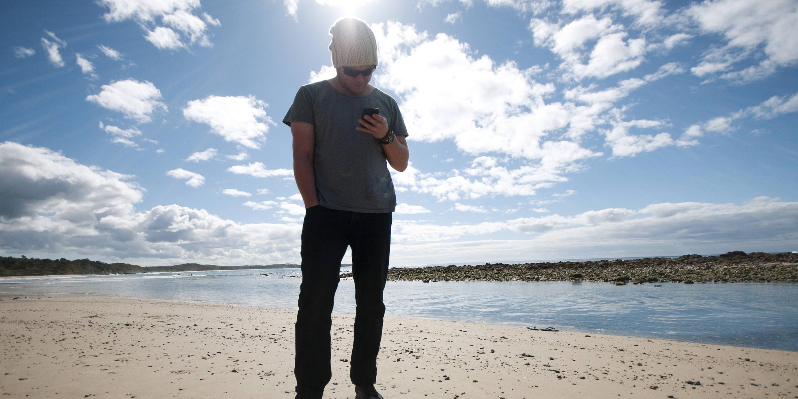 Mann steht am australischen Strand und schaut auf sein Handy 