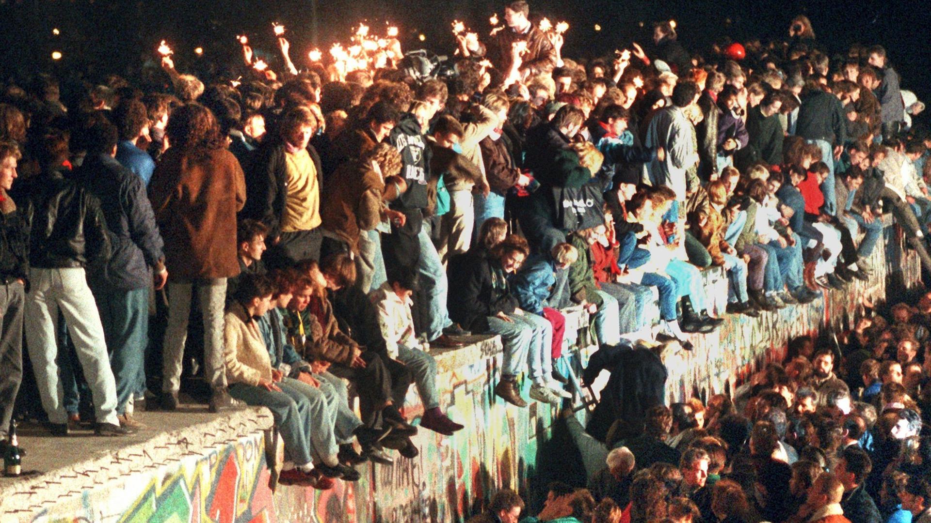 Zum Mauerfall in berlin 1989 sitzen und stehen viele Menschen jubelnd auf der Berliner Mauer.