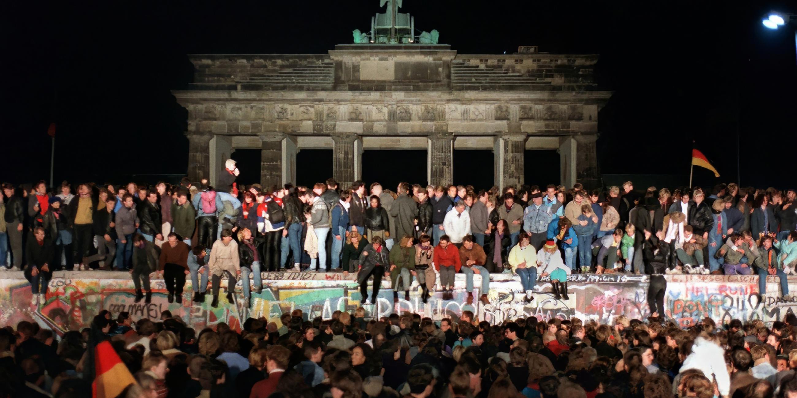 Auf der bunt bemalten Betonmauer vor dem Brandenburger Tor in Berlin sitzen und stehen Menschen. Davor hat sich eine Menschenmenge versammelt.