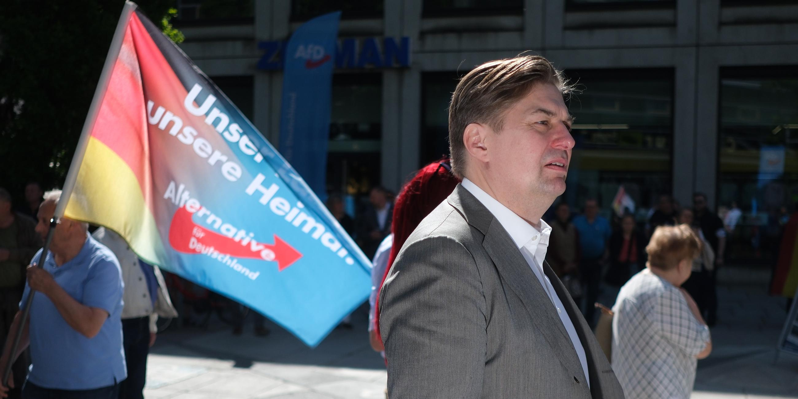 Ein Mann in Anzug läuft auf der Straße, im Hintergrund trägt ein Mann eine Flagge, auf der Unser Land, unsere Heimat und Alternative für Deutschland steht.