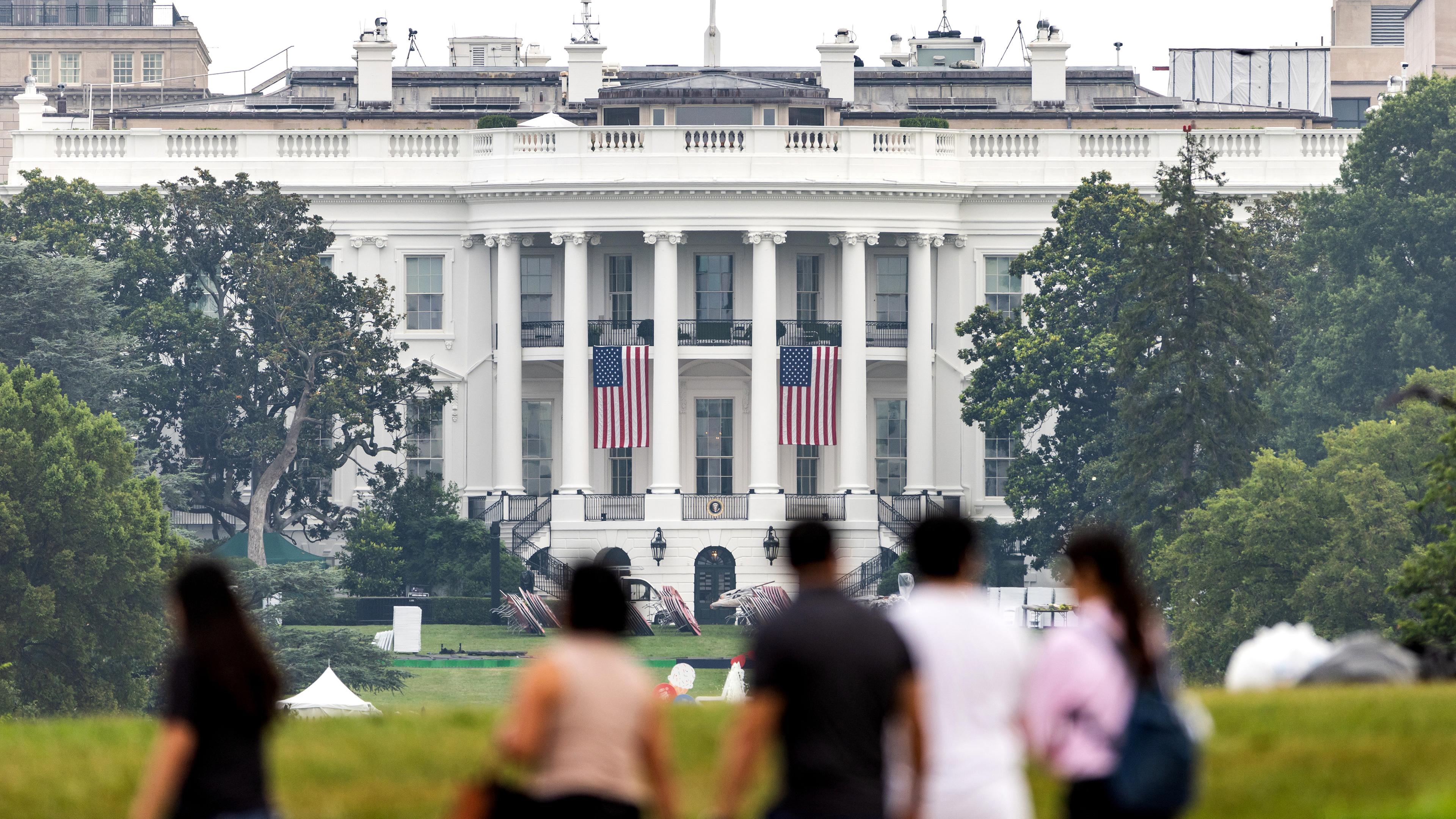Menschen gehen in Washington am Weißen Haus vorbei, dort hängen amerikanische Flaggen. 