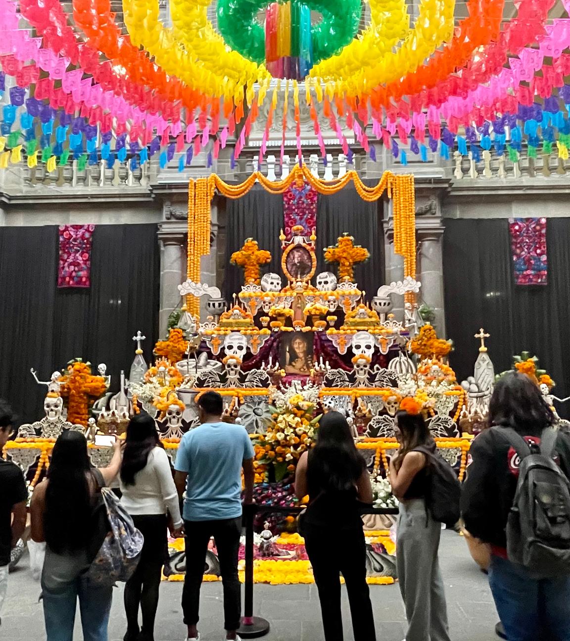 Ein Altar ist orange geschmückt, darauf stehen Skelettköpfe. Um ihn herum stehen Menschen.