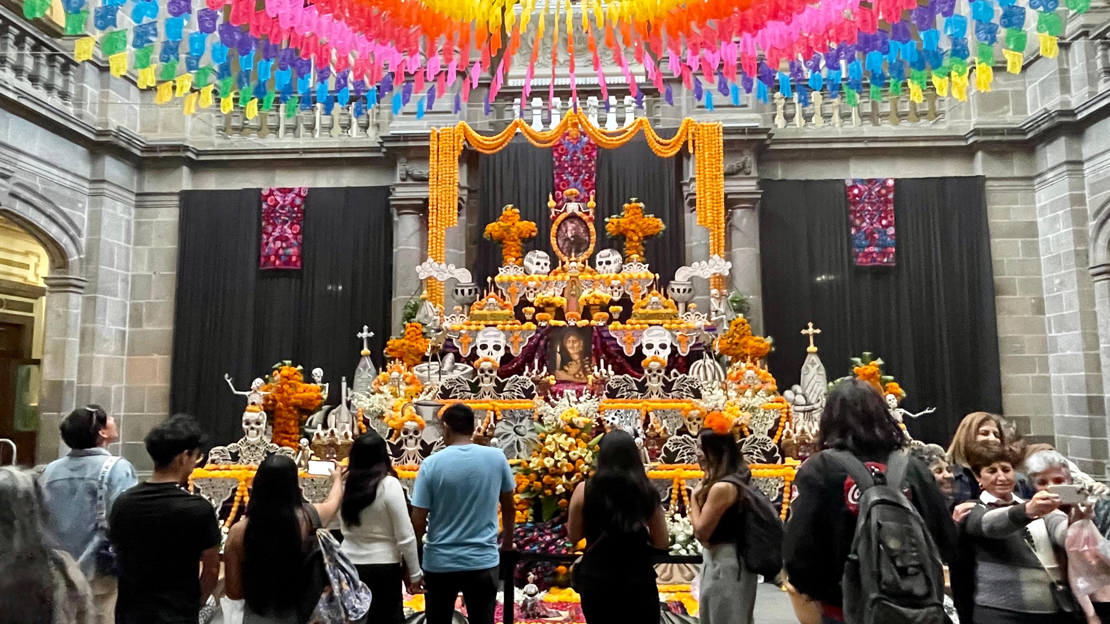 Ein Altar ist orange geschmückt, darauf stehen Skelettköpfe. Um ihn herum stehen Menschen.