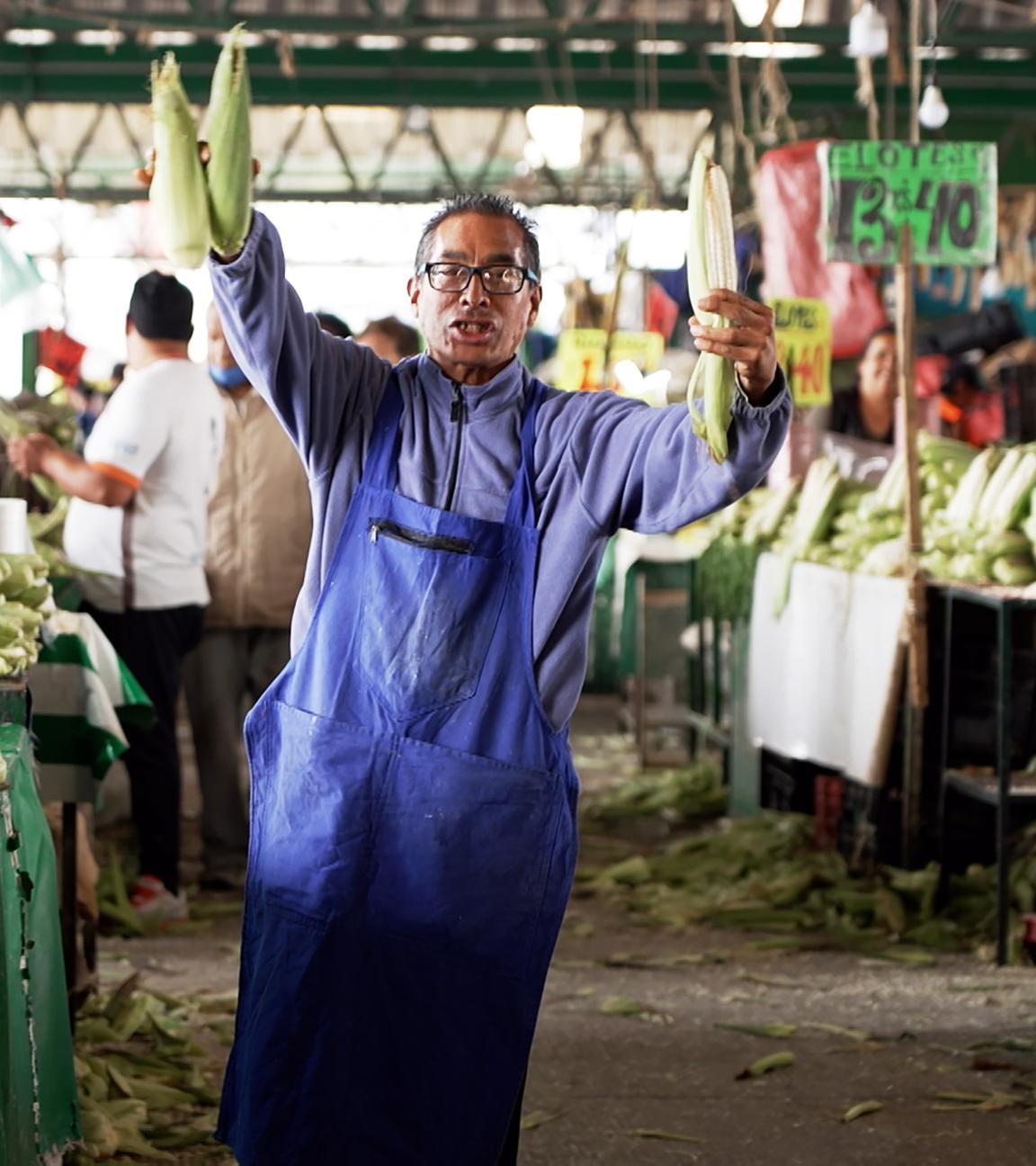 Auf einem Markt: Ein Mannes mittleren Alters trägt einen hellblauen Pullover und eine blaue Schürze. Mit beiden Händen hält er frischen Mais hoch. Um ihn herum stehen unzählige Obst- und Gemüsestände an denen einige Personen stehen.