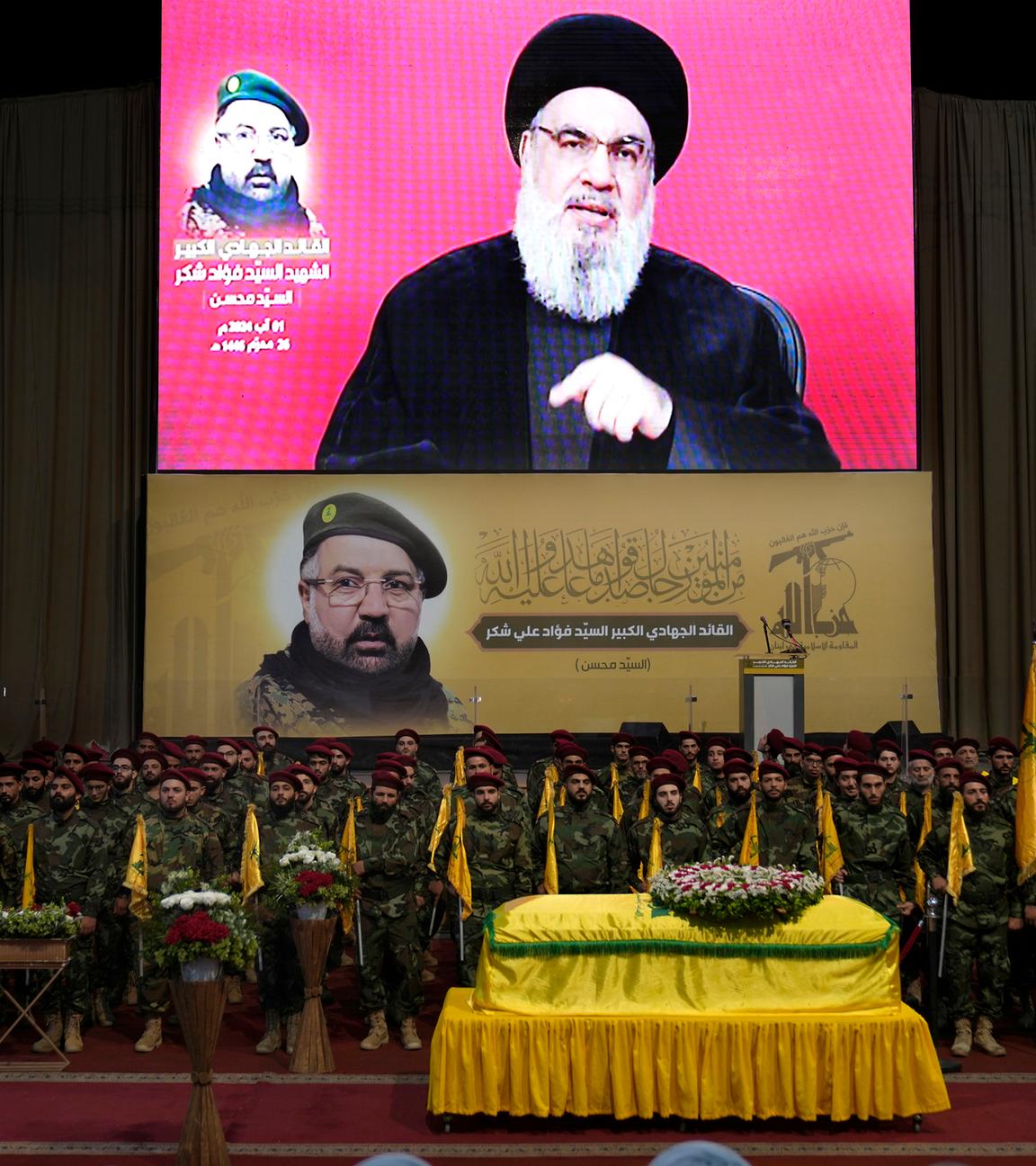 Libanon, Beirut: Hisbollah-Kämpfer tragen ihre Gruppenfahnen bei einem Trauerzug, während sie hinter dem Sarg ihres Befehlshabers Fouad Shukur stehen. Hassan Nasrallah, Hisbollah-Generalsekretär, auf einem Bildschirm zu sehen