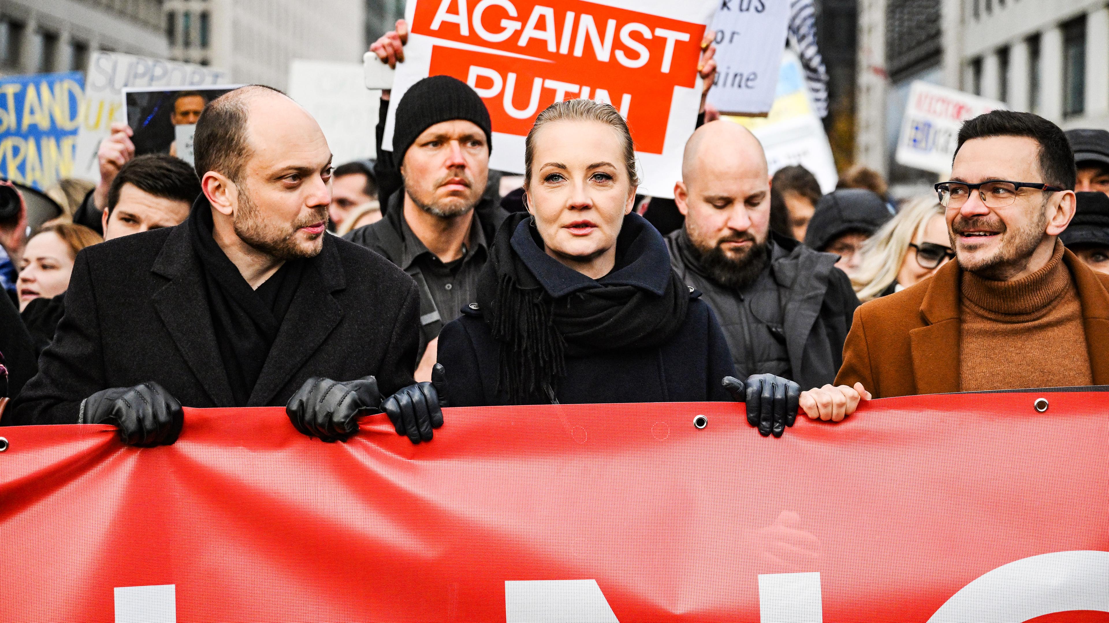 Eine Frau mit streng zurückgekämmten Haaren und bekleidet mit dicker Winterjacke und Schal läuft lächelnd bei einer Demo mit. Hinter ihr hält eine Frau ein rotes Schild mit der Aufschrift Against Putin hoch.