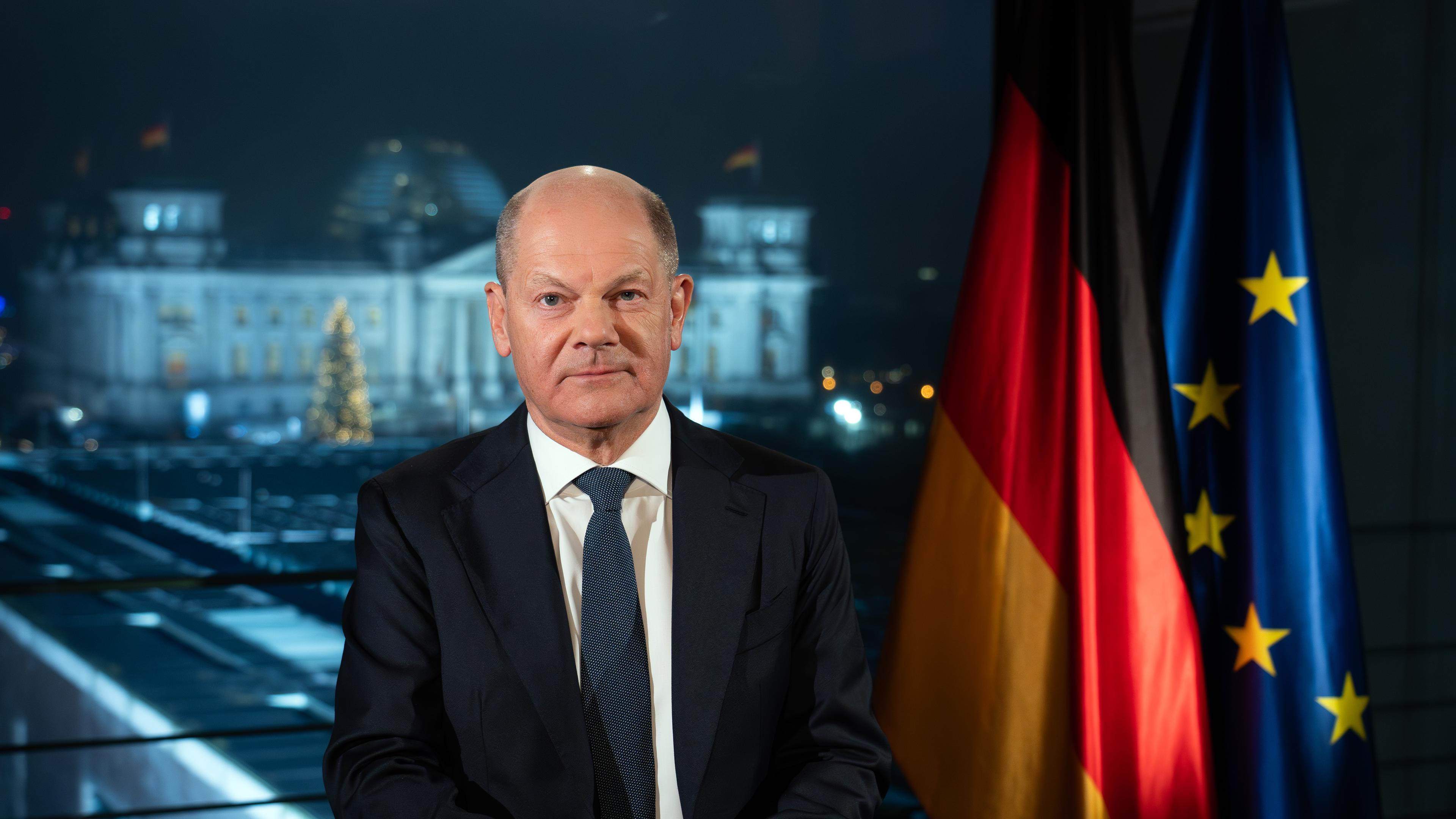 Bundeskanzler Olaf Scholz (SPD) sitzt während eines Fototermins nach der Aufzeichnung seiner Neujahrsansprache im Kanzleramt vor der Kulisse des von Scheinwerfern angestrahlten Reichstages.