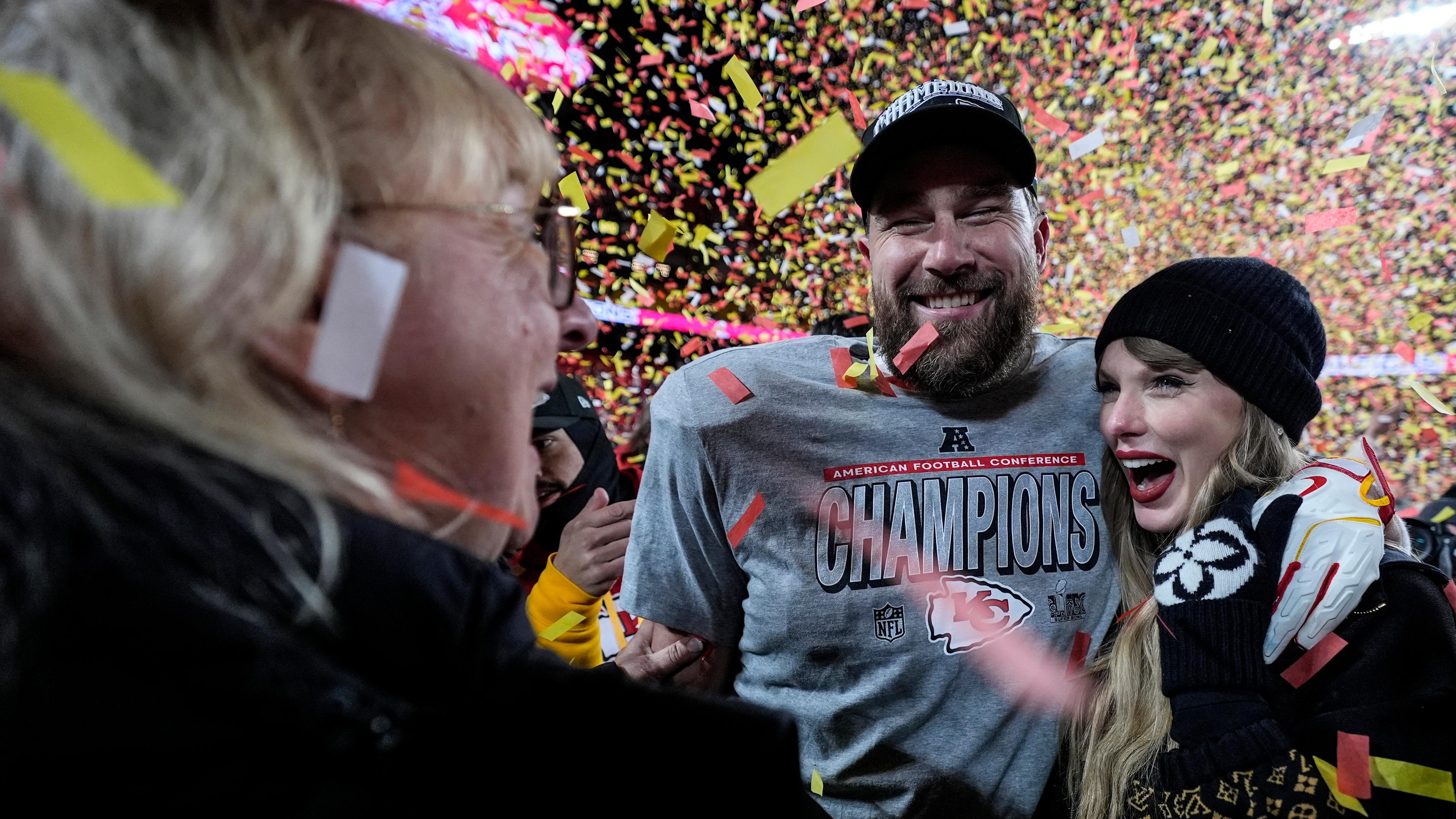 Taylor Swift und Travis Kelce stehen Arm in Arm in einem gelb-roten Konfettiregen und lachen mit breit geöffneten Mündern. Kelce trägt eine schwarze Kappe und ein graues Football-Trikot mit der Aufschrift "Champions". Swift trägt roten Lippenstift und eine schwarze Mütze.  