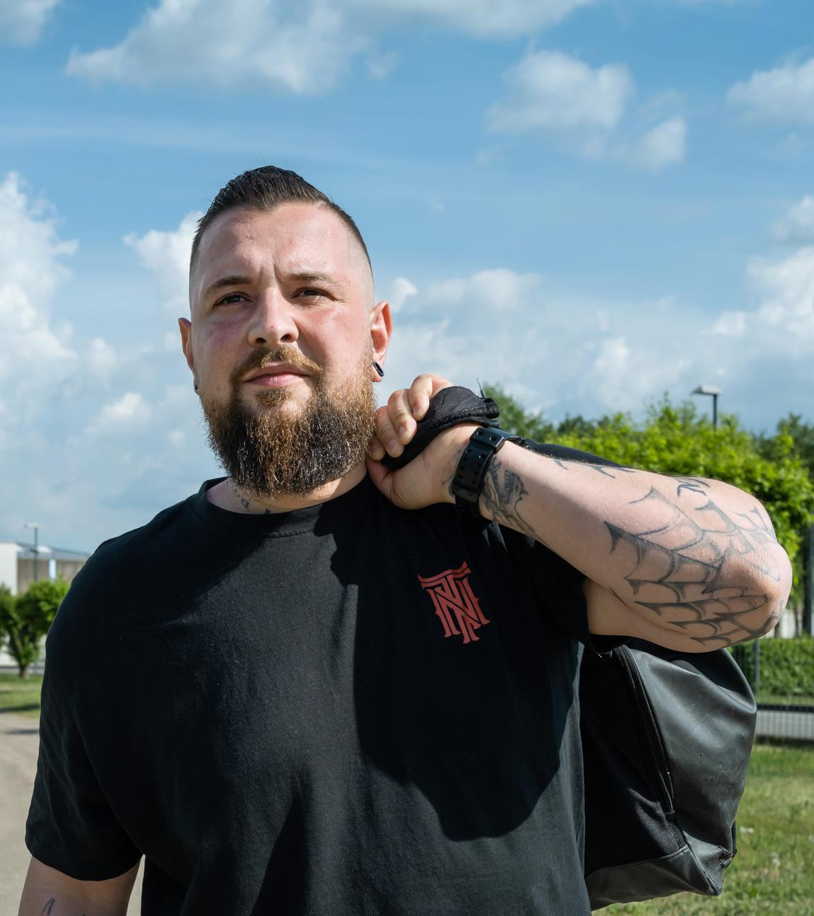 Alex (32) läuft auf die Kamera zu. Er trägt ein schwarzes T-Shirt und eine schwarze Hose. Sein linker Arm ist angewinkelt, er trägt eine Reisetasche um seine linke Schulter. Er geht einen Weg entlang, der von niedriges Gras gesäumt ist. Im Hintergrund steht ein niedriges Gefängnisgebäude aus grauem Beton mit blauem Tor.