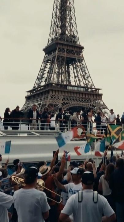Menschen schwenken Flaggen vor einem Boot auf der Seine; im Hintergrund der Eiffelturm in Paris.