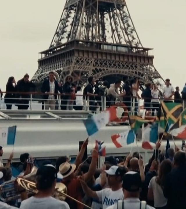 Menschen schwenken Flaggen vor einem Boot auf der Seine; im Hintergrund der Eiffelturm in Paris.