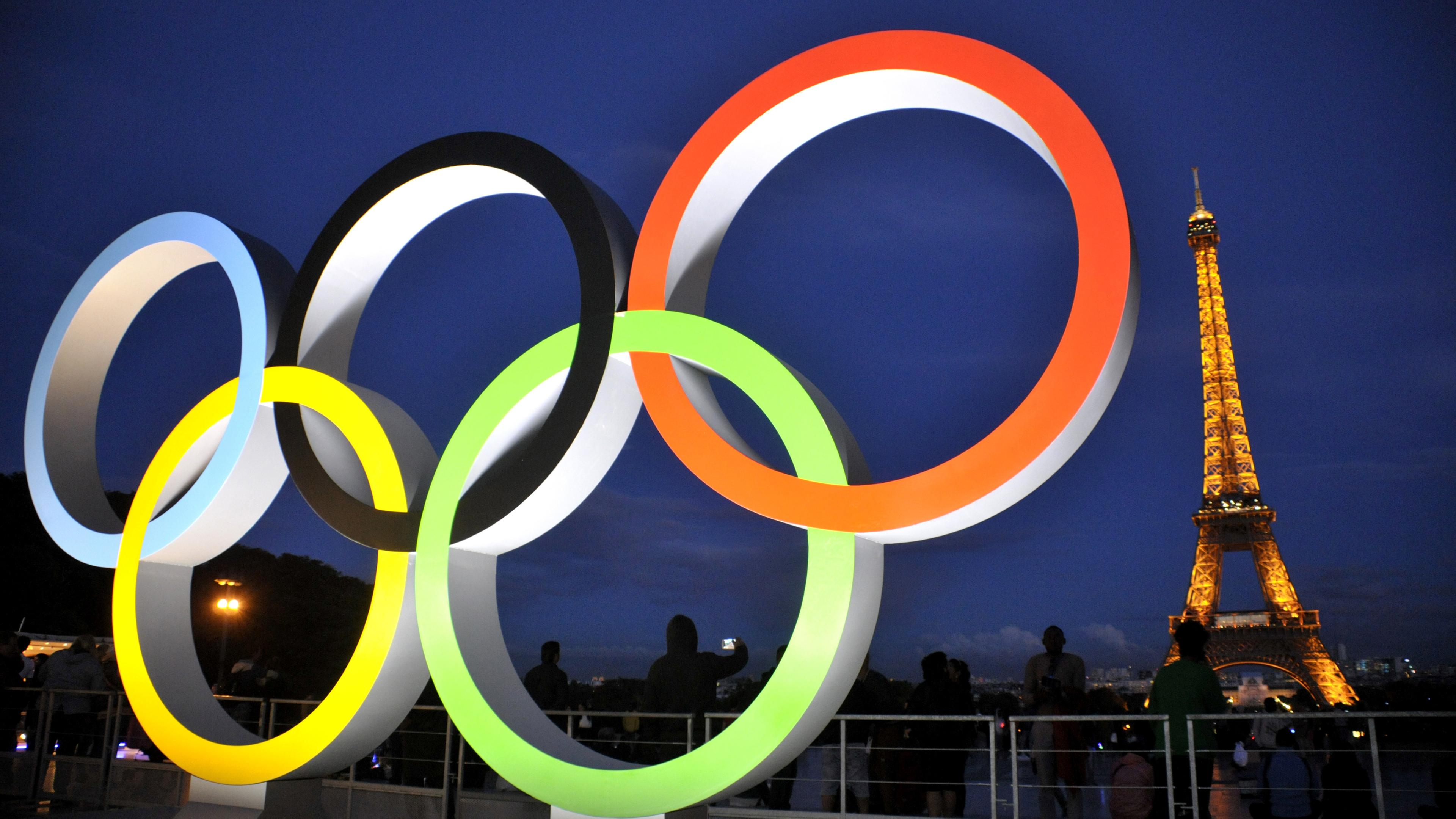 Die beleuchteten olympischen Ringe im Vordergrund. Im Hintergrund der Eiffelturm beleuchtet.
