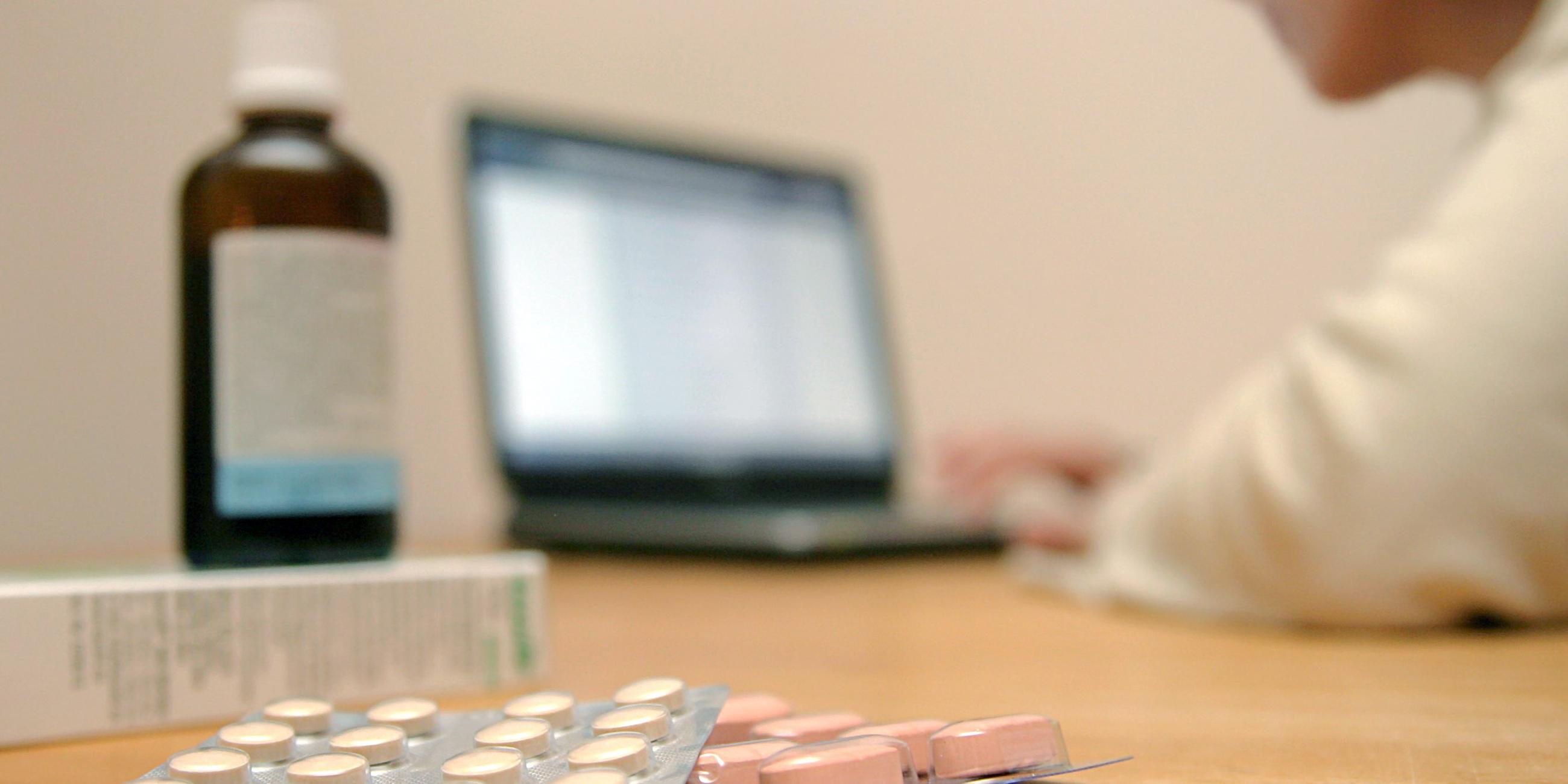 Eine Person sitzt vor einem Laptop und bestellt in einer Onlineapotheke. Neben ihr stehen einige Medikamente.