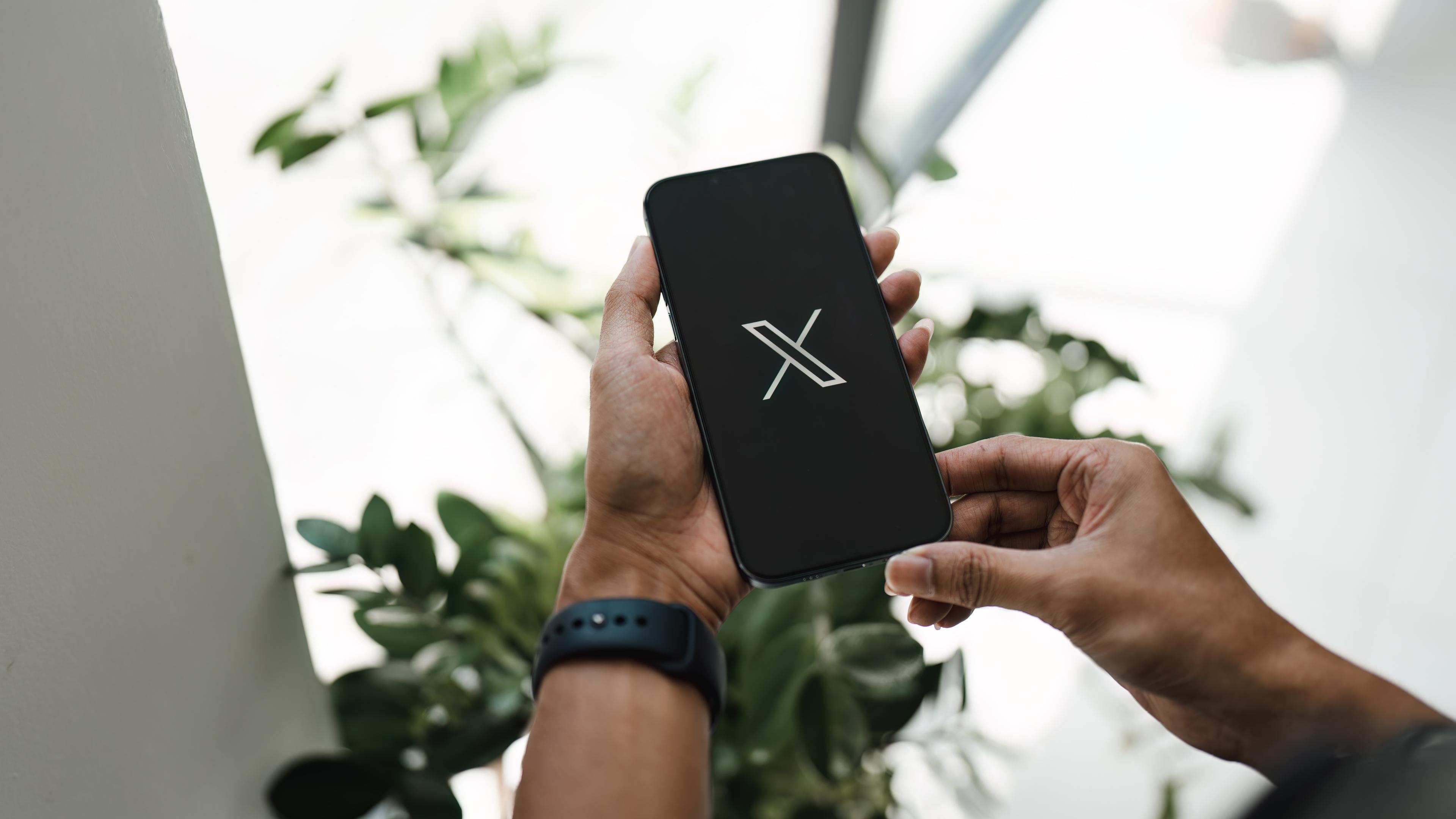 Eine Person hält ein Smartphone in der Hand. Auf dem Bildschirm ist das Logo der Plattform X zu sehen.