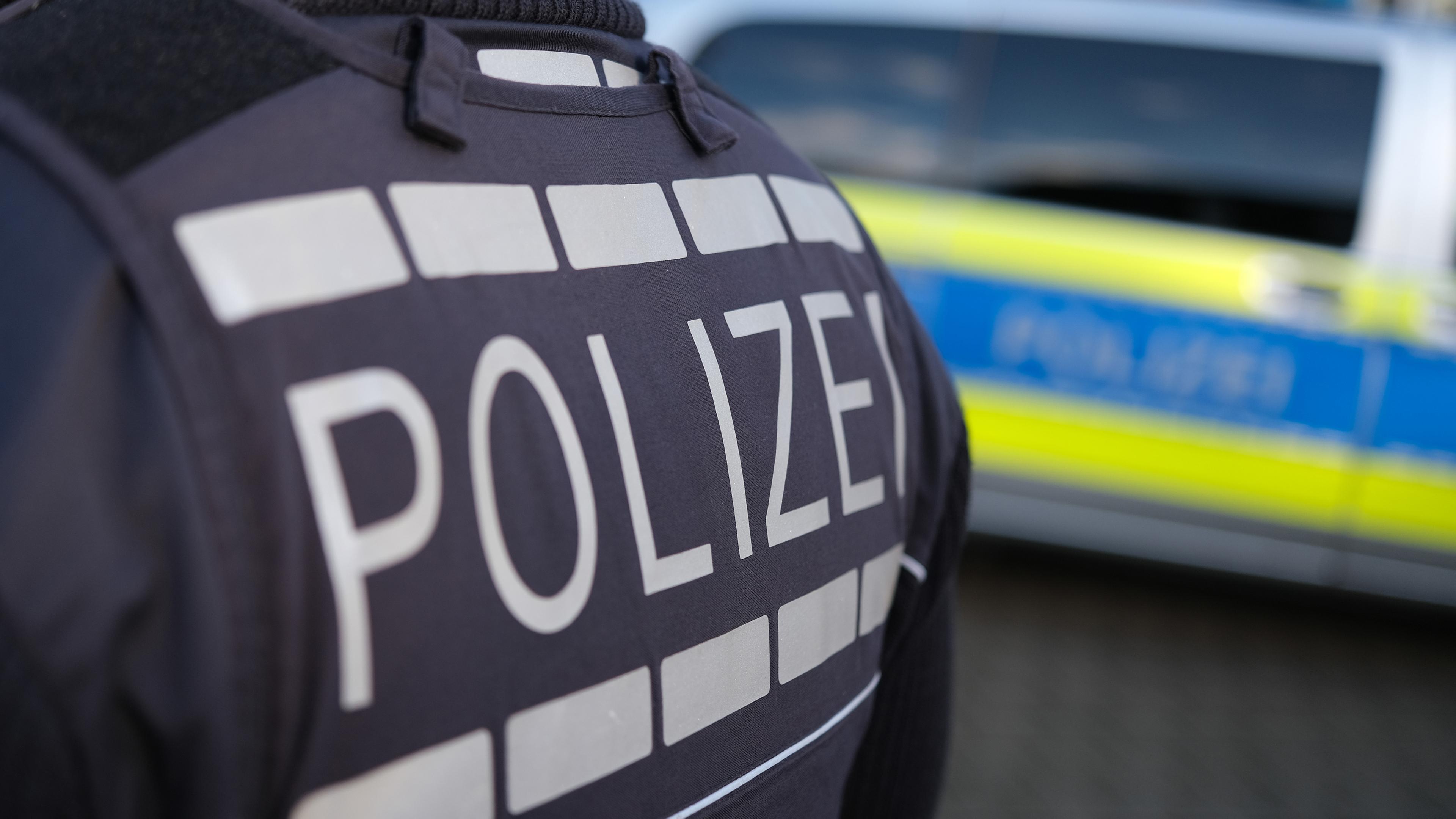 Rücken eines Polizisten mit Aufschrift Polizei, im Hintergrund ein Polizeiauto (Symbolbild)