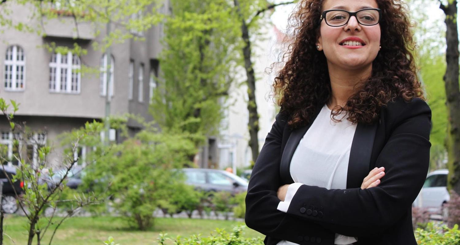 Eine Frau mit dunklen Haaren und Brille steht mit verschränkten Armen in einem Park. Sie lächelt freundlich.