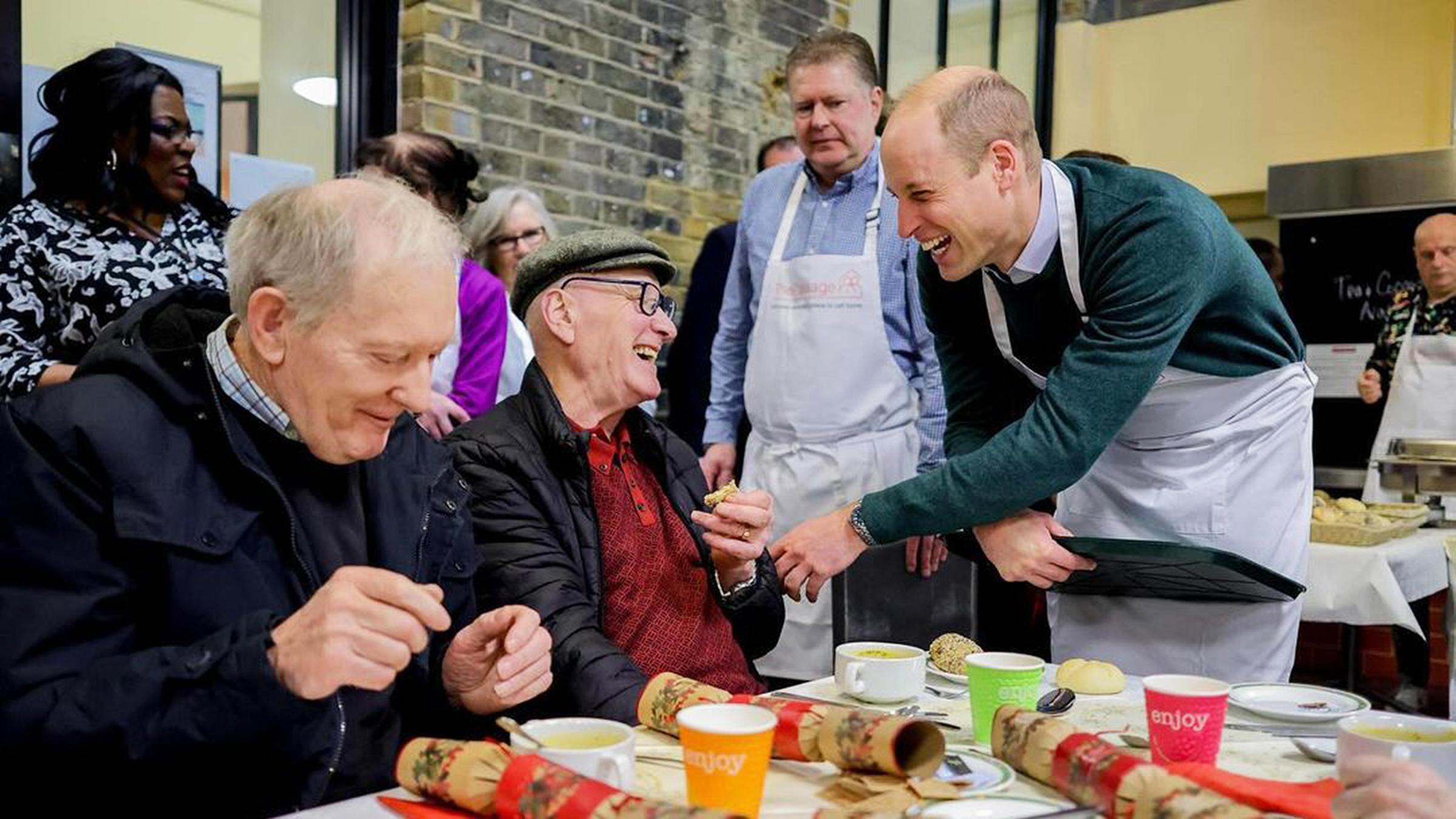 Prinz William in Kochschürze lacht mit einem älteren Mann.