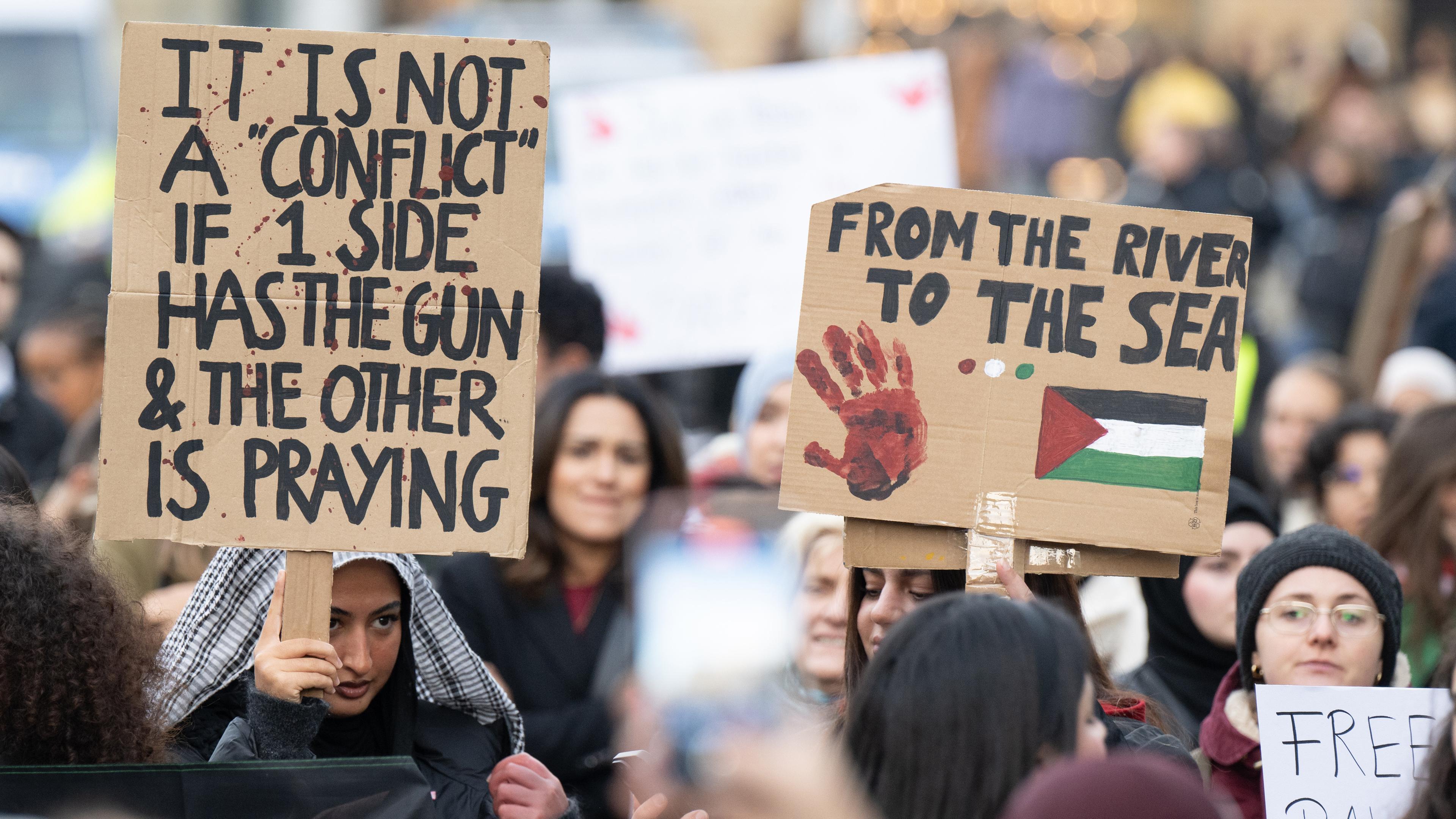 Hessen, Frankfurt/Main: Junge Frauen halten Plakate mit Aufschriften "It is not a "conflict" if 1 side has the gun & the other is praying" und "From the river to the sea" bei einer pro-palästinensischen Demonstration in Frankfurt hoch.