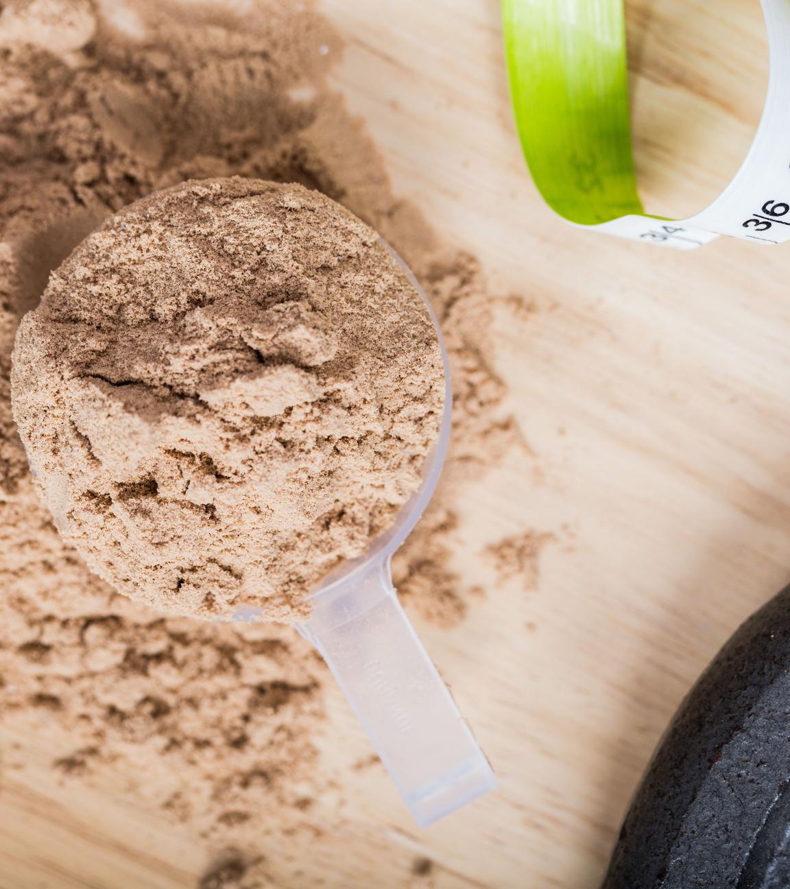 Ein Messbecher mit Molkenproteinpulver liegt auf einem Holztisch neben einem Maßband und einem Gewicht.