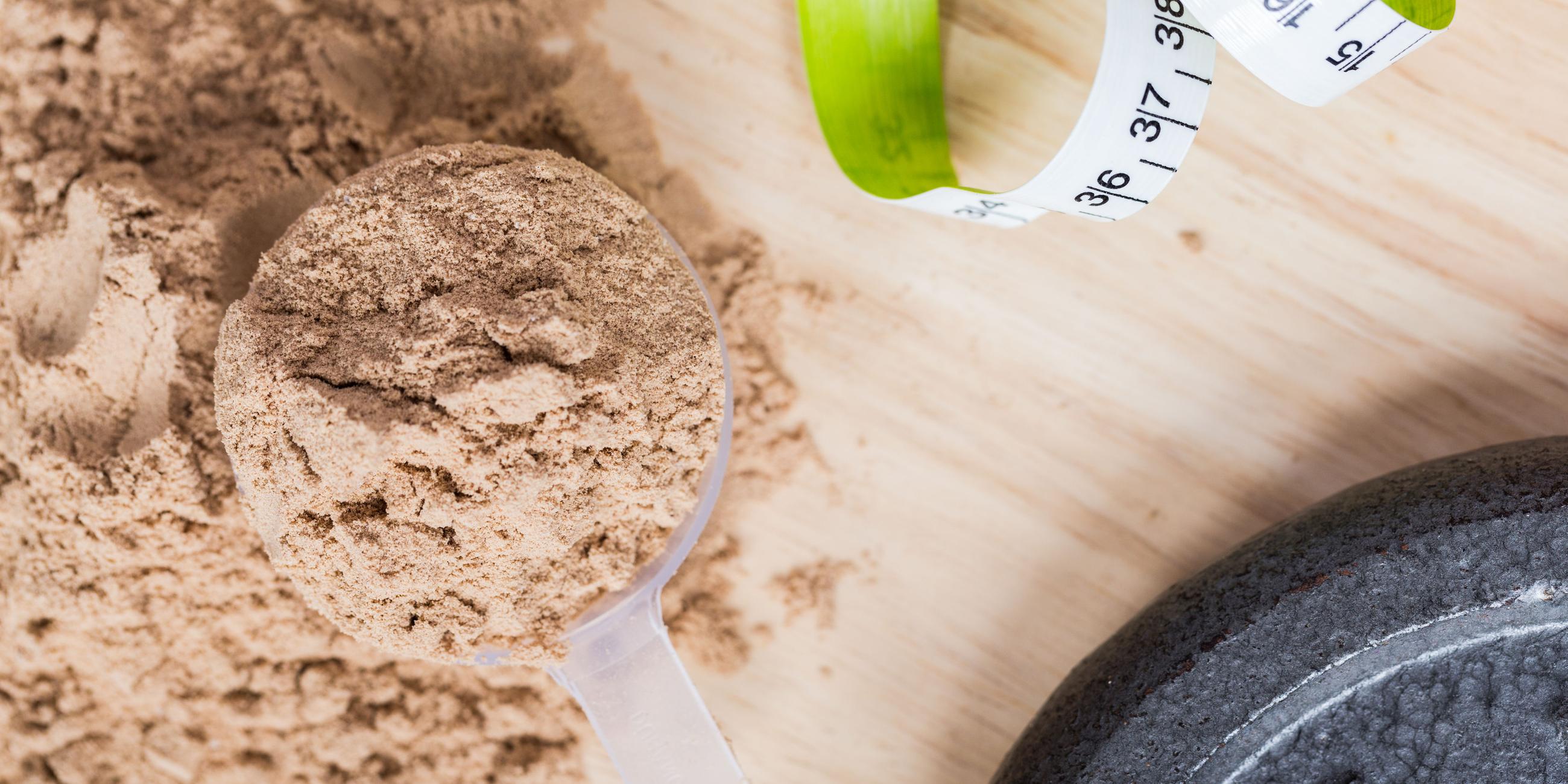 Ein Messbecher mit Molkenproteinpulver liegt auf einem Holztisch neben einem Maßband und einem Gewicht.