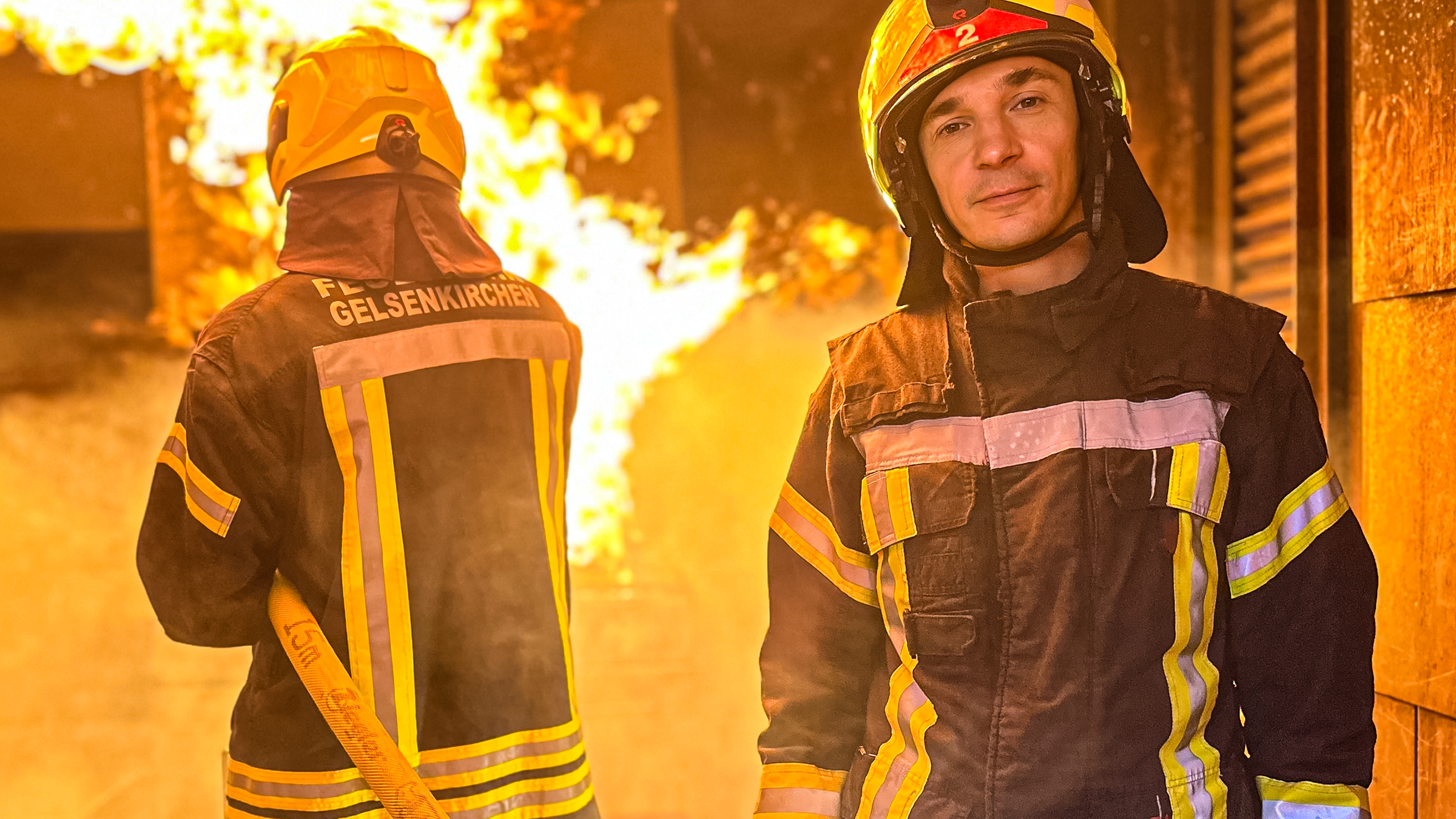 PUR+ Moderator Eric Mayer steht in Feuerwehr-Montur und mit Helm vor einem Feuer. Ein anderer Feuerwehrmann, der nur von hintenzu sehen ist, löscht mit einem Schlauch das Feuer.