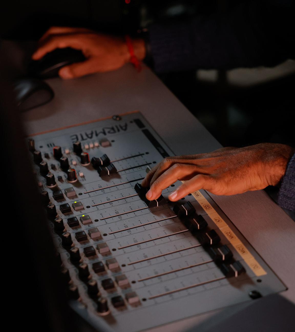 Eine Person sitzt in einem Tonstudio. Nur die Hände sind zu sehen: die eine hält eine Maus, die andere schiebt einen Regler im Tonstudio