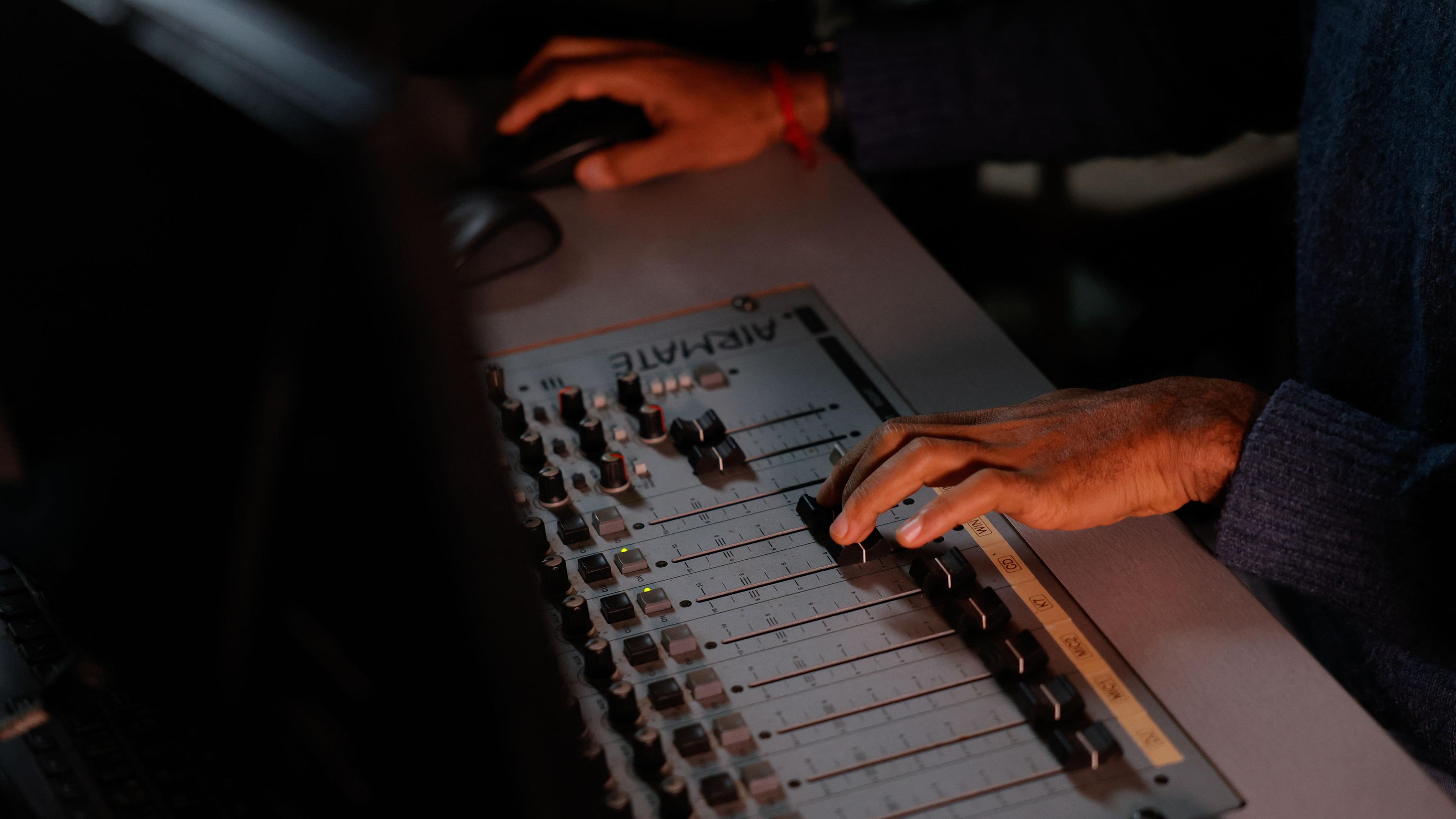 Eine Person sitzt in einem Tonstudio. Nur die Hände sind zu sehen: die eine hält eine Maus, die andere schiebt einen Regler im Tonstudio