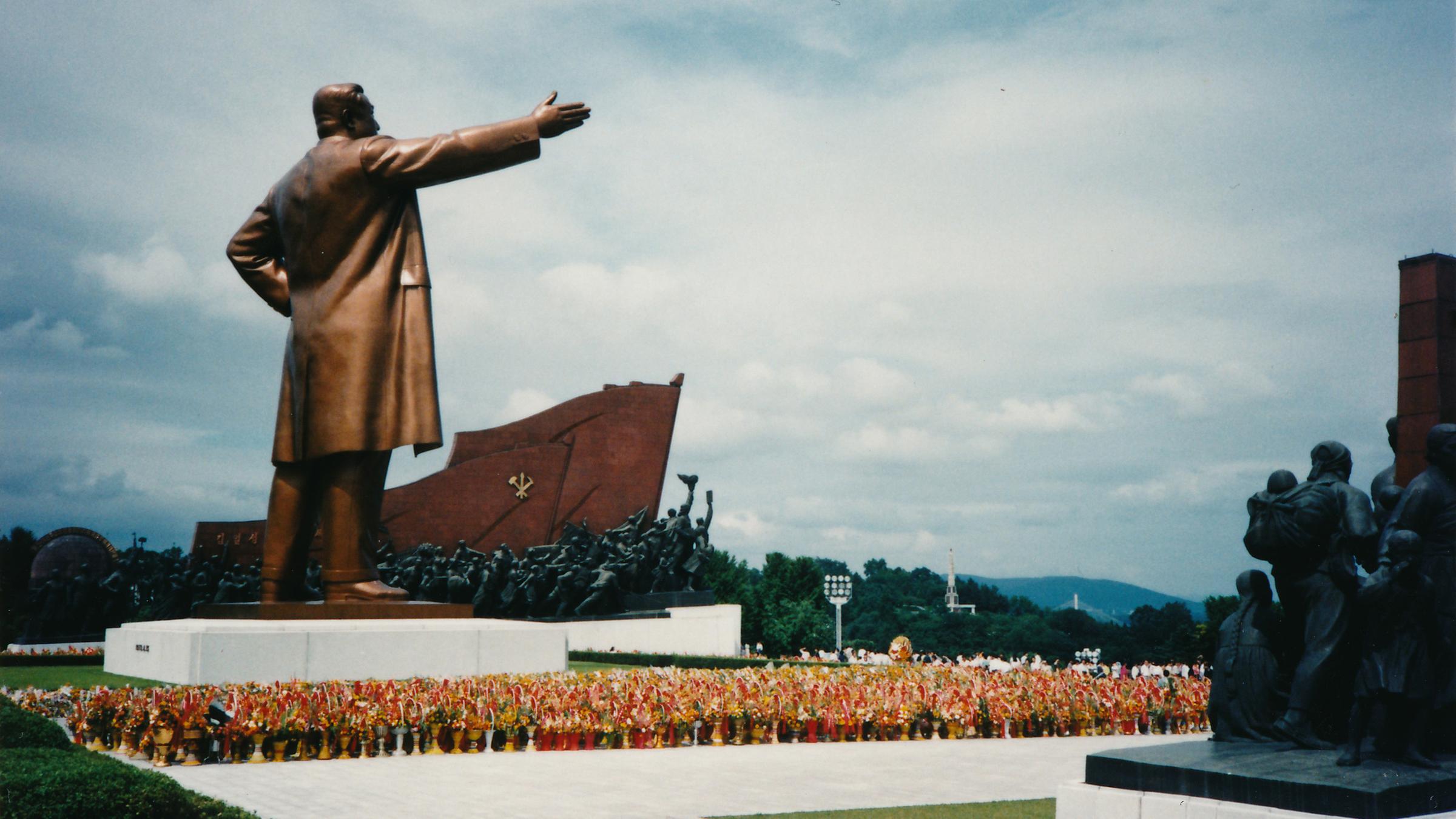 Auf einem Platz steht eine große, bronzefarbene Statue. Vor ihr sind viele Menschen in roter und gelber Uniform versammelt.