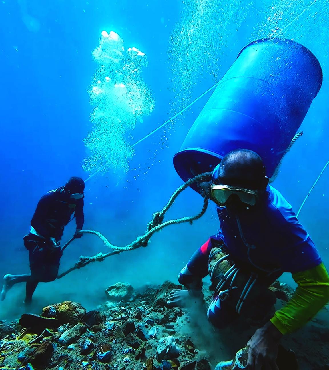 Am Meeresgrund: Zwei Taucher bestücken ein Netz mit goldhaltigem Gestein. Eine Kunststofftonne schwimmt zwischen ihnen. Diese wird dann mit Luft gefüllt. Der Auftrieb bringt das Gestein dann an die Wasseroberfläche.