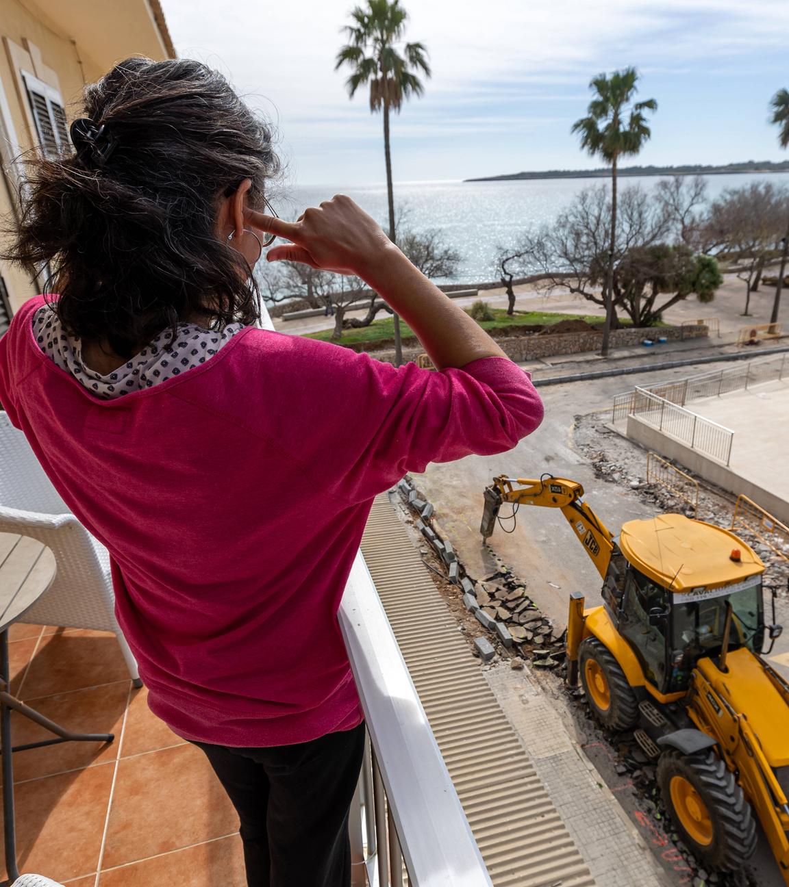 Eine Frau sitzt auf dem Balkon ihres Hotels und hält sich die Ohren zu, während ein Bagger auf der Baustelle vor dem Hotel arbeitet.