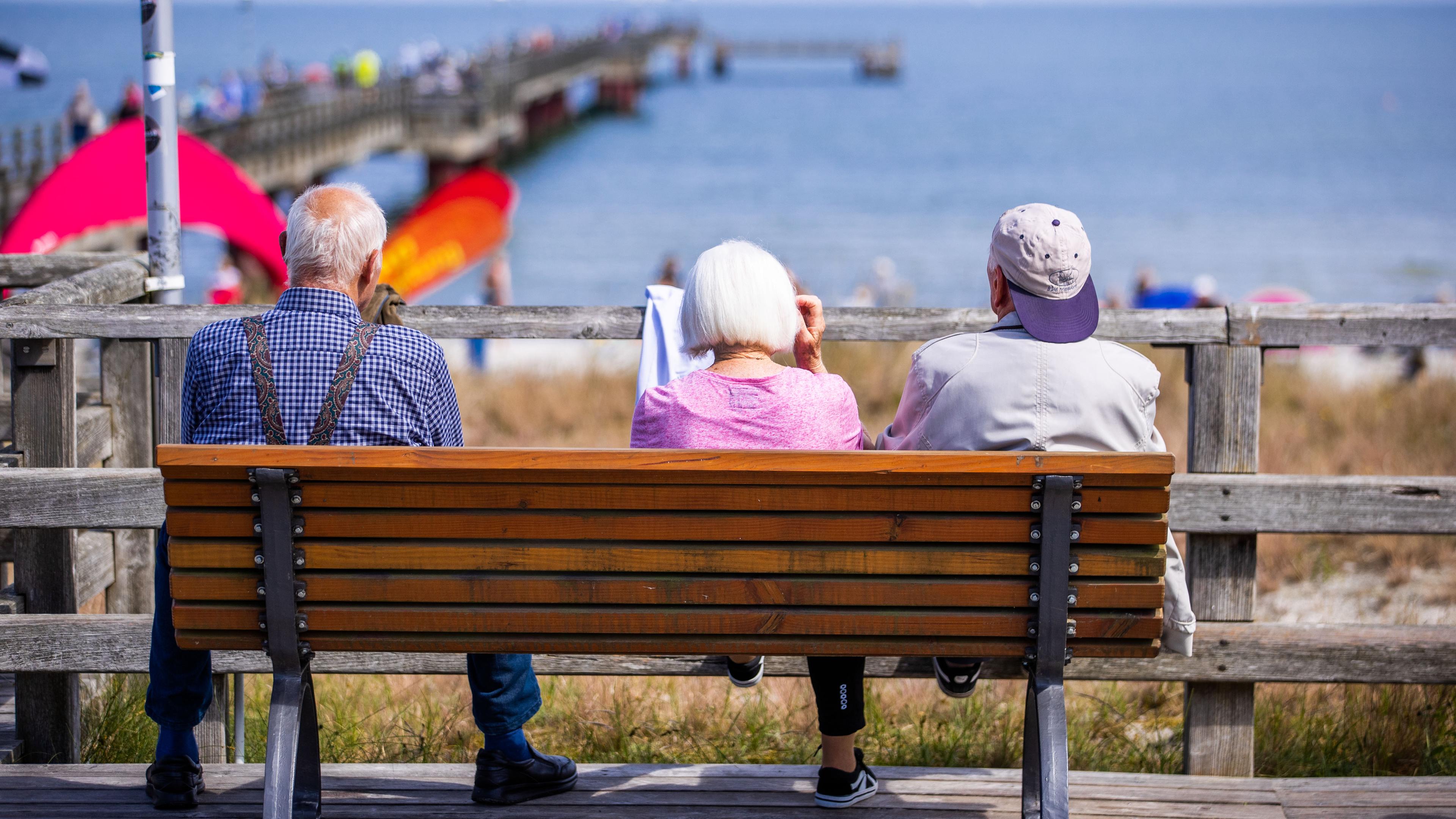Drei ältere Urlauber sitzen auf einer Bank an der Seebrücke.