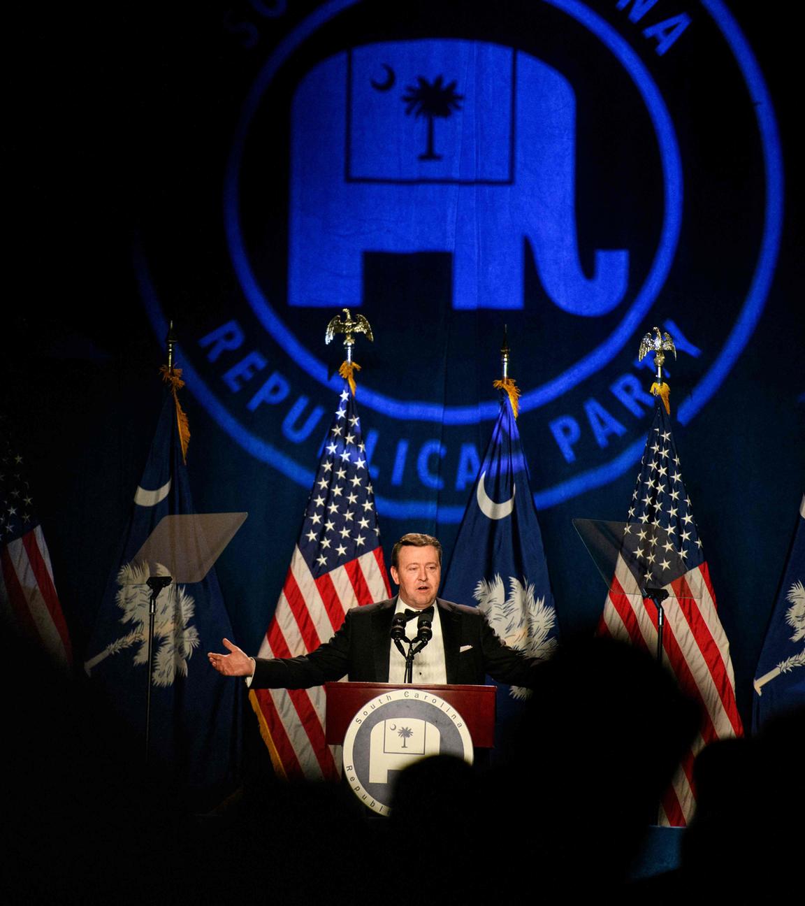 Ein Mann steht auf einer Bühne und spricht an einem Pult, vor ihm sind amerikanische Flaggen und im Hintergrund wird das Symbol der republikanischen Partei (der Elefant) angestrahlt.