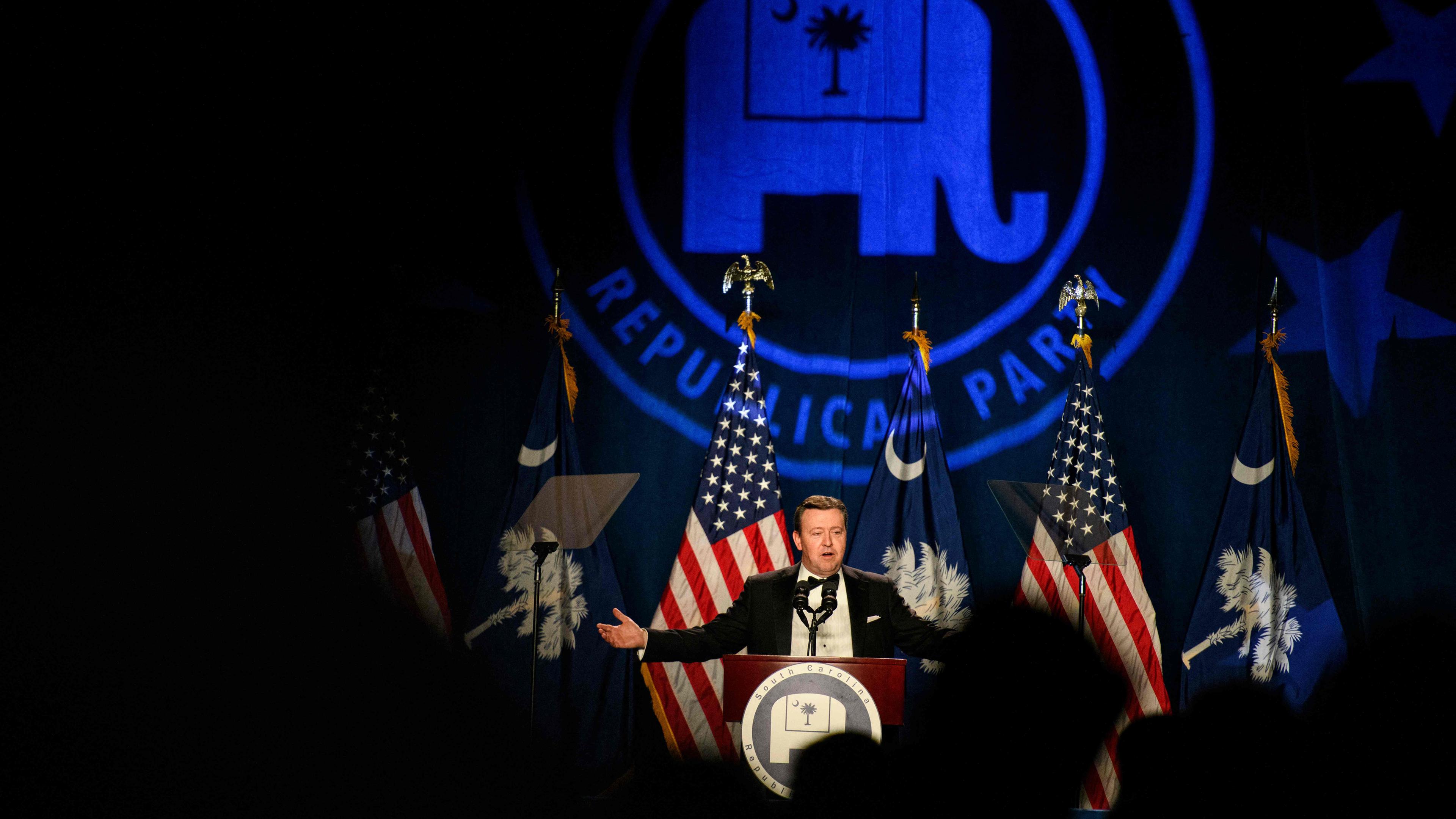 Ein Mann steht auf einer Bühne und spricht an einem Pult, vor ihm sind amerikanische Flaggen und im Hintergrund wird das Symbol der republikanischen Partei (der Elefant) angestrahlt.