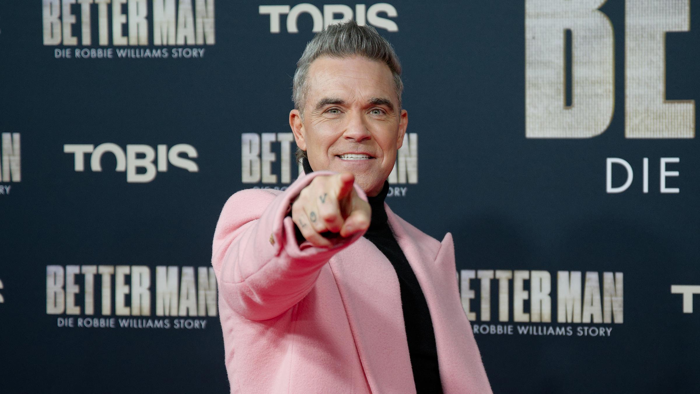 Im rosafarbenen Mantel war Robbie Williams der Hingucker bei der Premiere “Better Man“ in Köln. Ein computergenerierter Affe spielt in seiner Filmbiografie den Ex-Take-That-Star. 