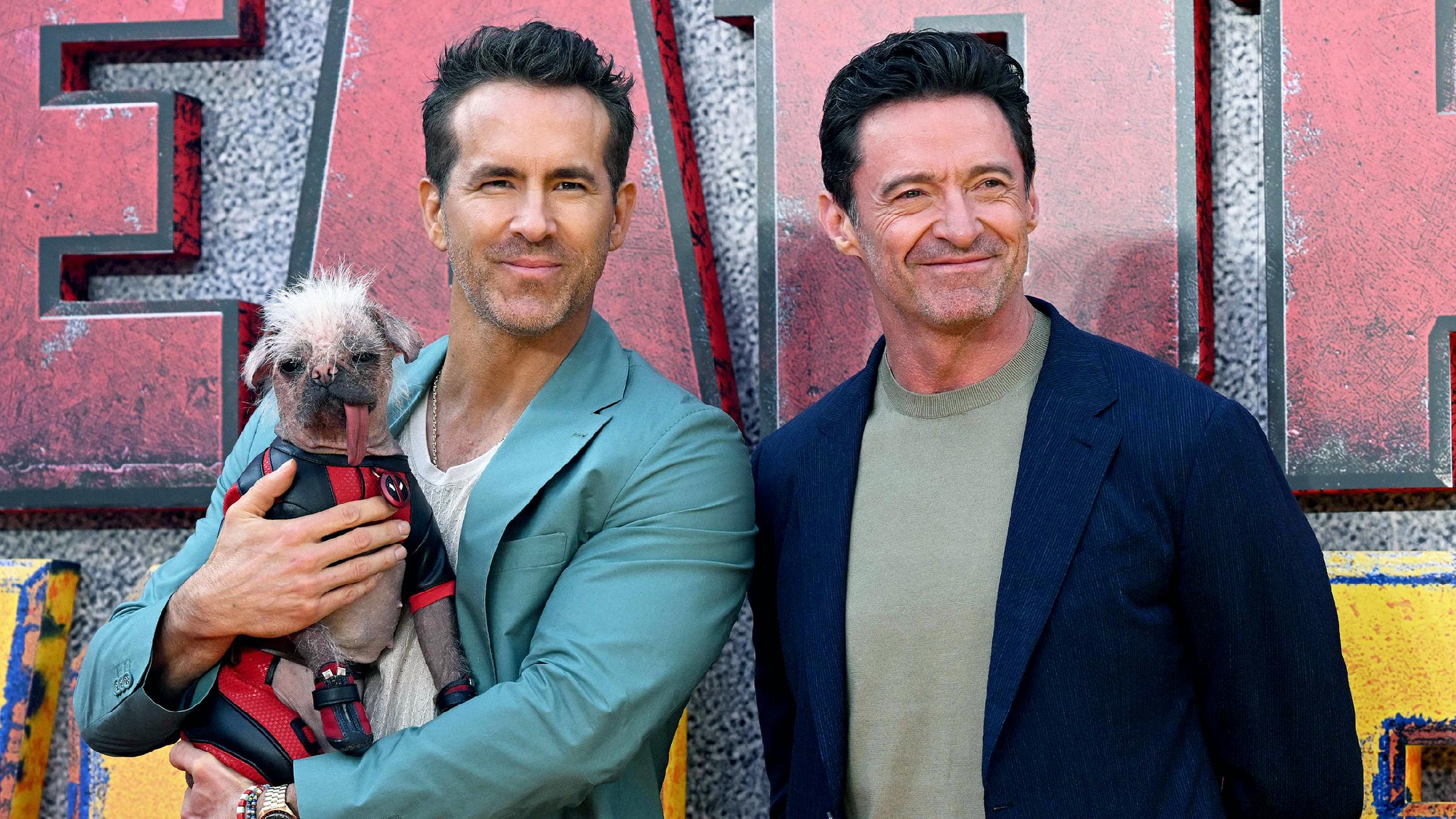 Hugh Jackman steht neben Ryan Reynolds, dieser hält Hund Peggy im Arm.