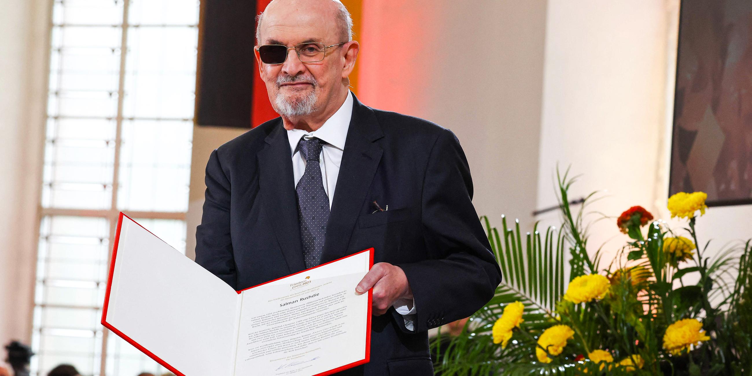 Ein älterer Mann mit Glatze und graumeliertem Bart trägt eine Brille deren eines Glas schwarz verdunkelt ist und einen Anzug mit Krawatte. In der Hand hält er eine Mappe mit einer Preisurkunde.